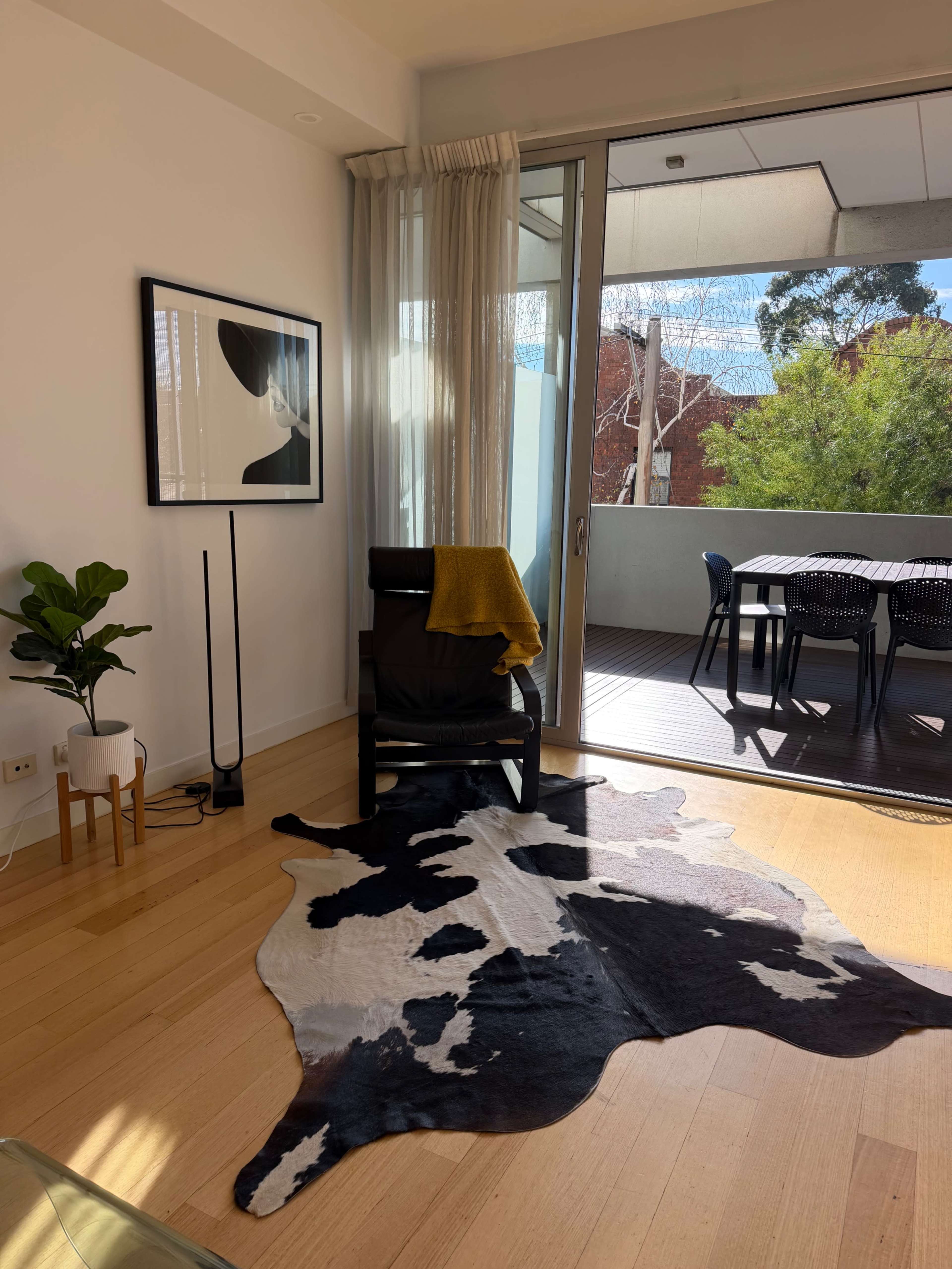 The image shows a modern living room with a black and white cowhide rug, a chair draped with a yellow throw, and large glass doors leading to a balcony.