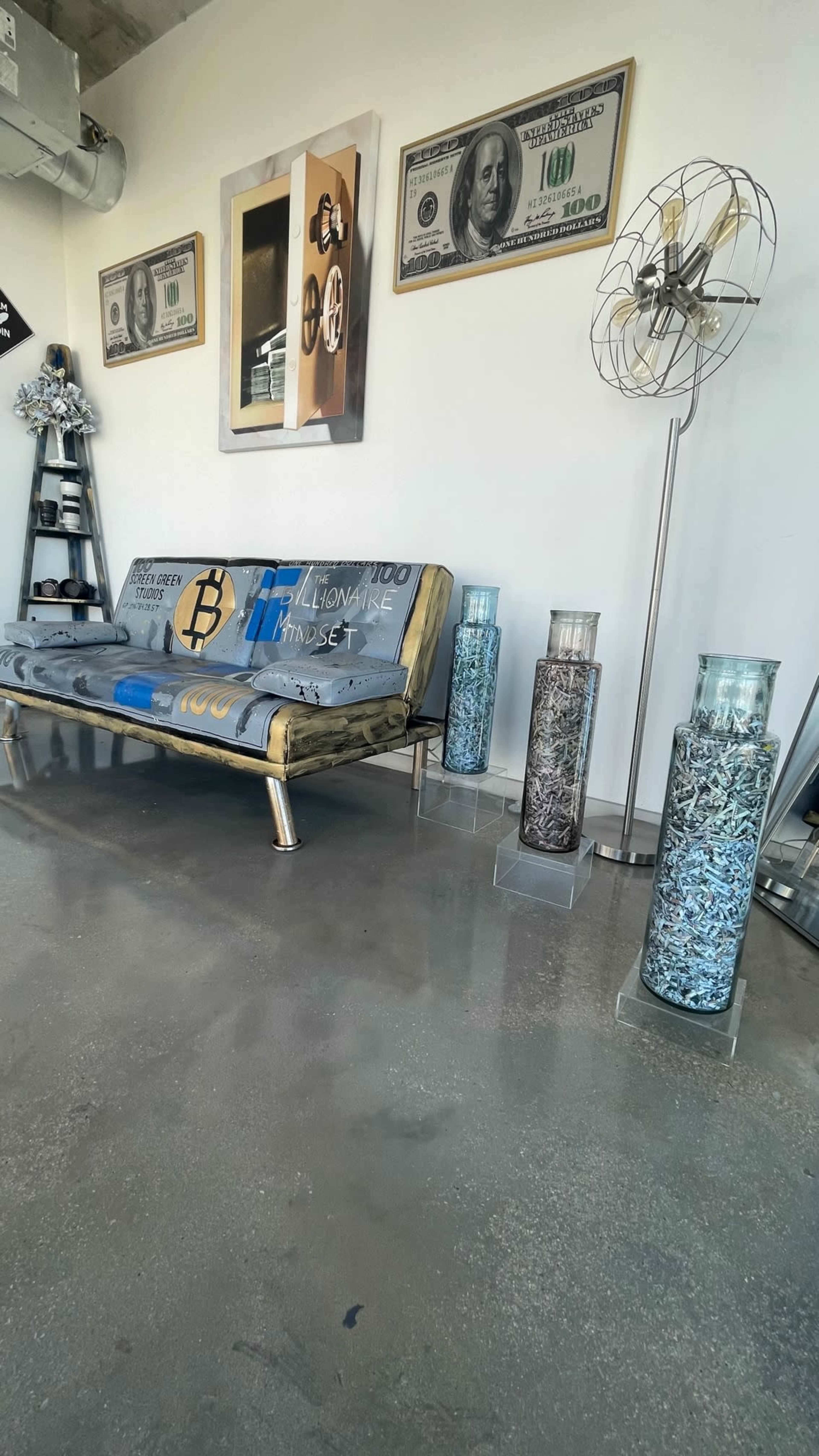 A modern interior with a blue couch, framed artwork, large dollar bill prints, and two glass containers filled with small objects on a polished concrete floor.