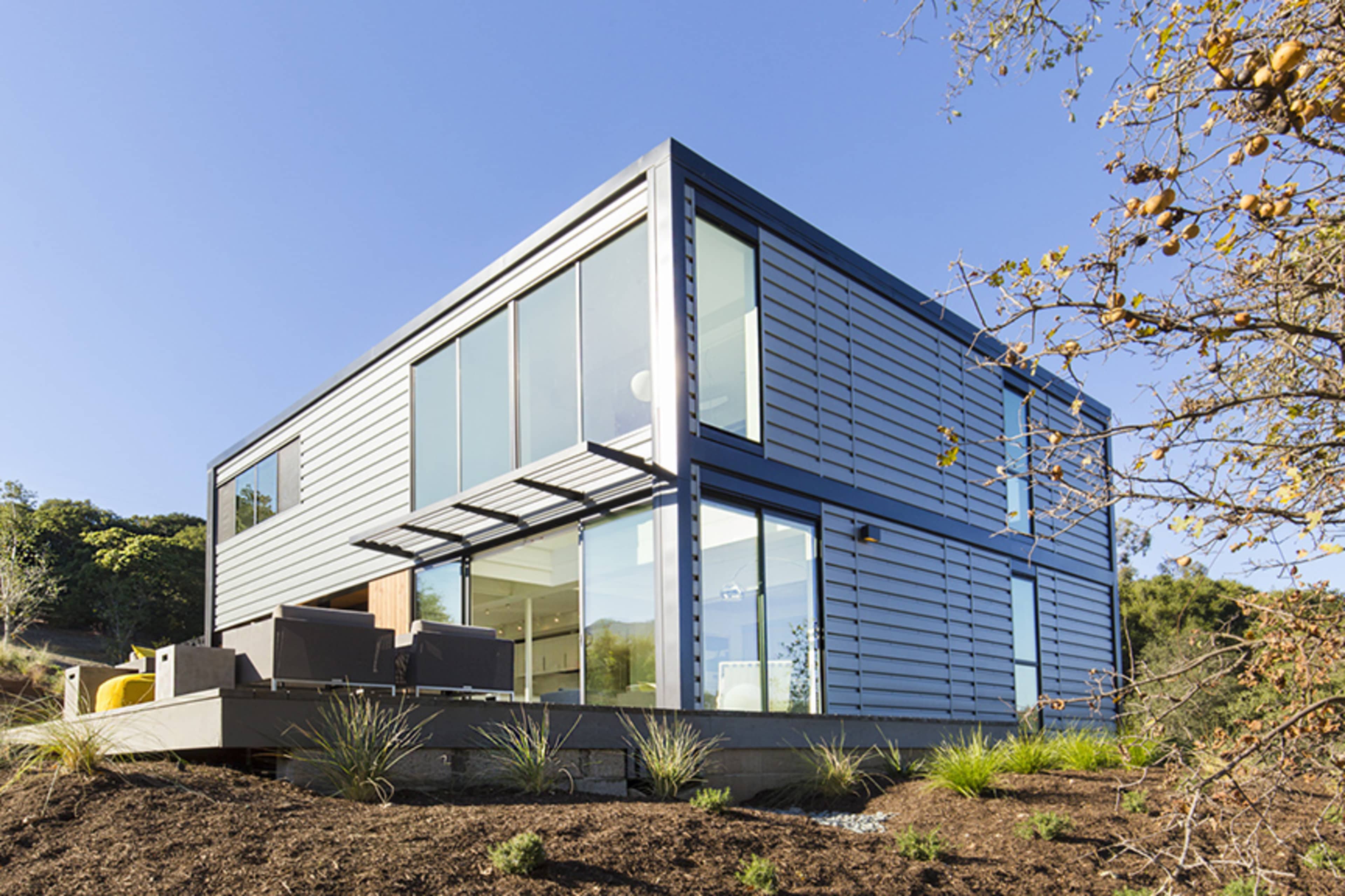 A modern, two-story house with a metal exterior and large windows is situated on a hillside surrounded by landscaping.