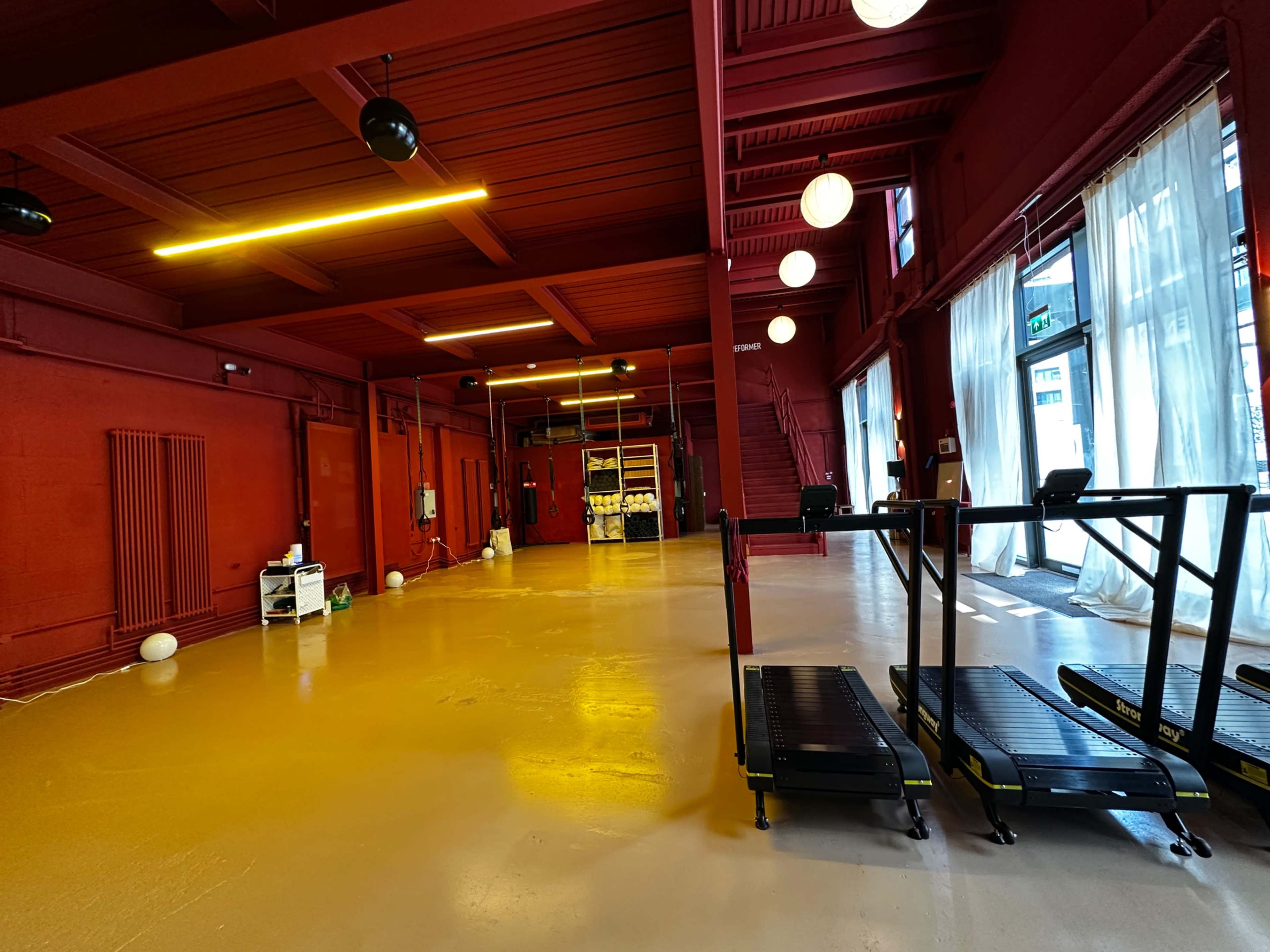 The image shows a spacious, red-painted gym area with two treadmill machines and large windows allowing natural light to enter.