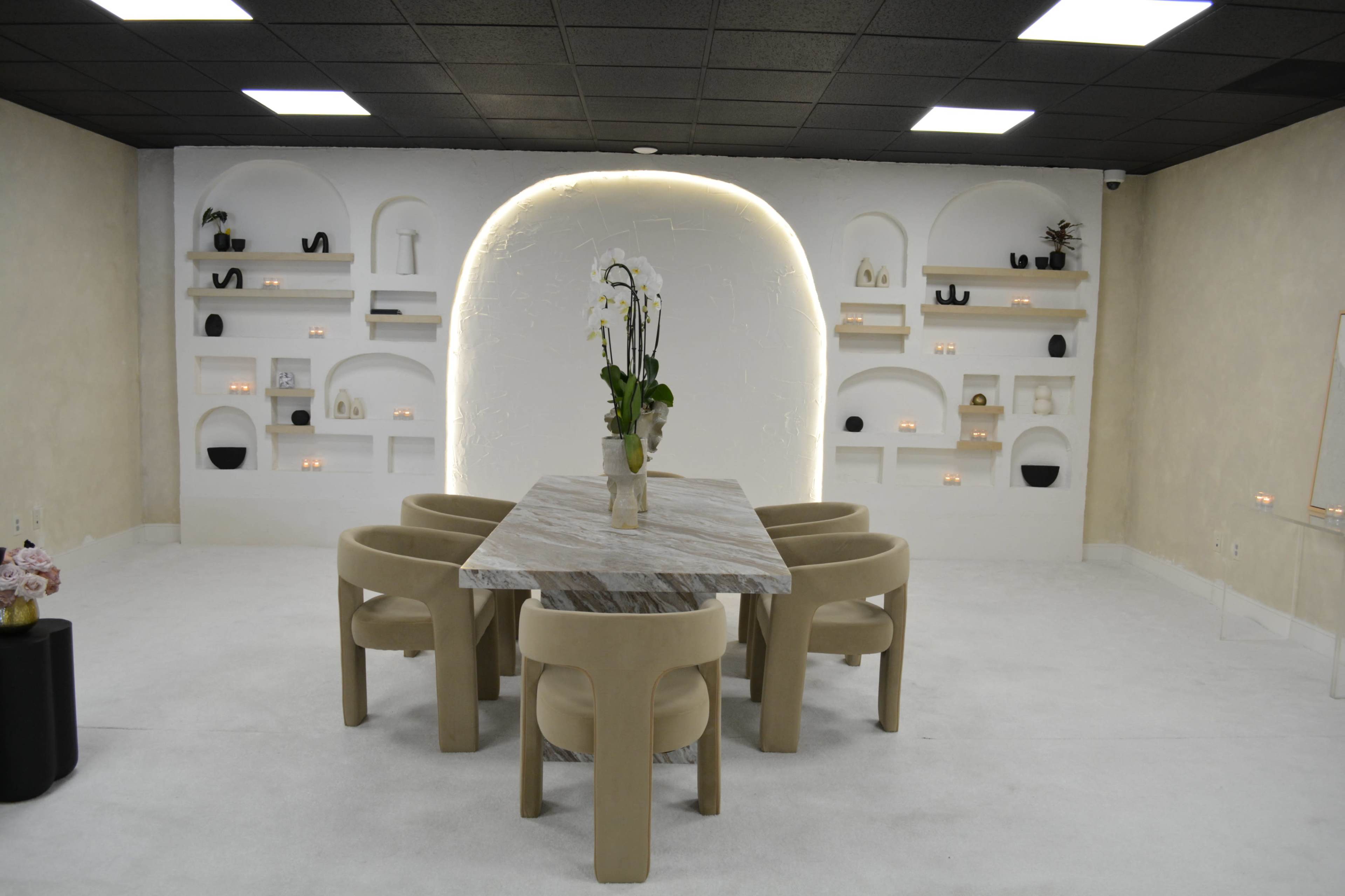 A modern dining space features a marble table surrounded by four beige chairs, with an illuminated arch and a decorative wall shelf in the background.