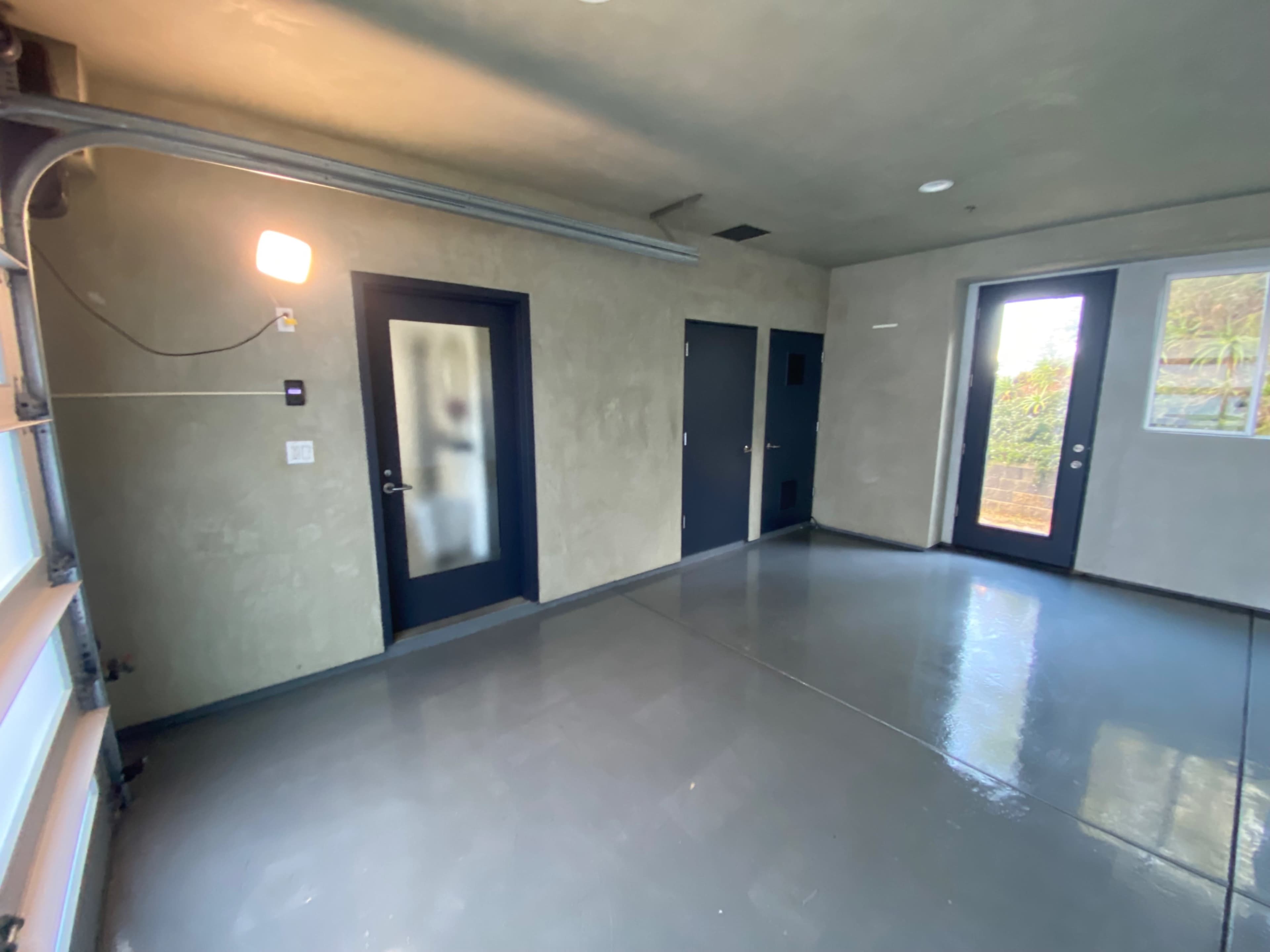 The image shows a minimalist, empty room with concrete walls, a polished floor, and two doors leading outside.