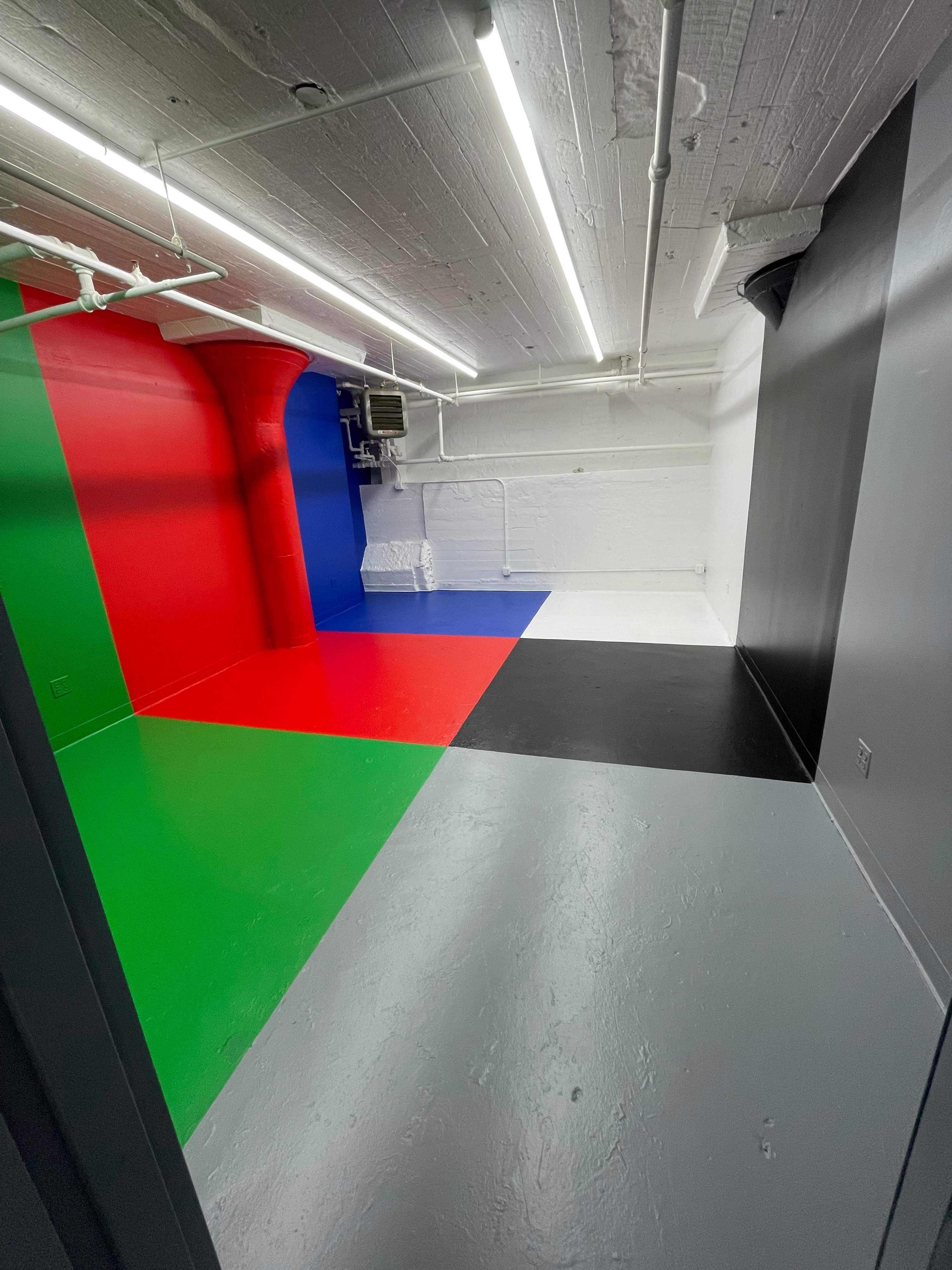 A room with a multi-colored floor divided into green, red, blue, and black sections, with white walls and exposed pipes on the ceiling.
