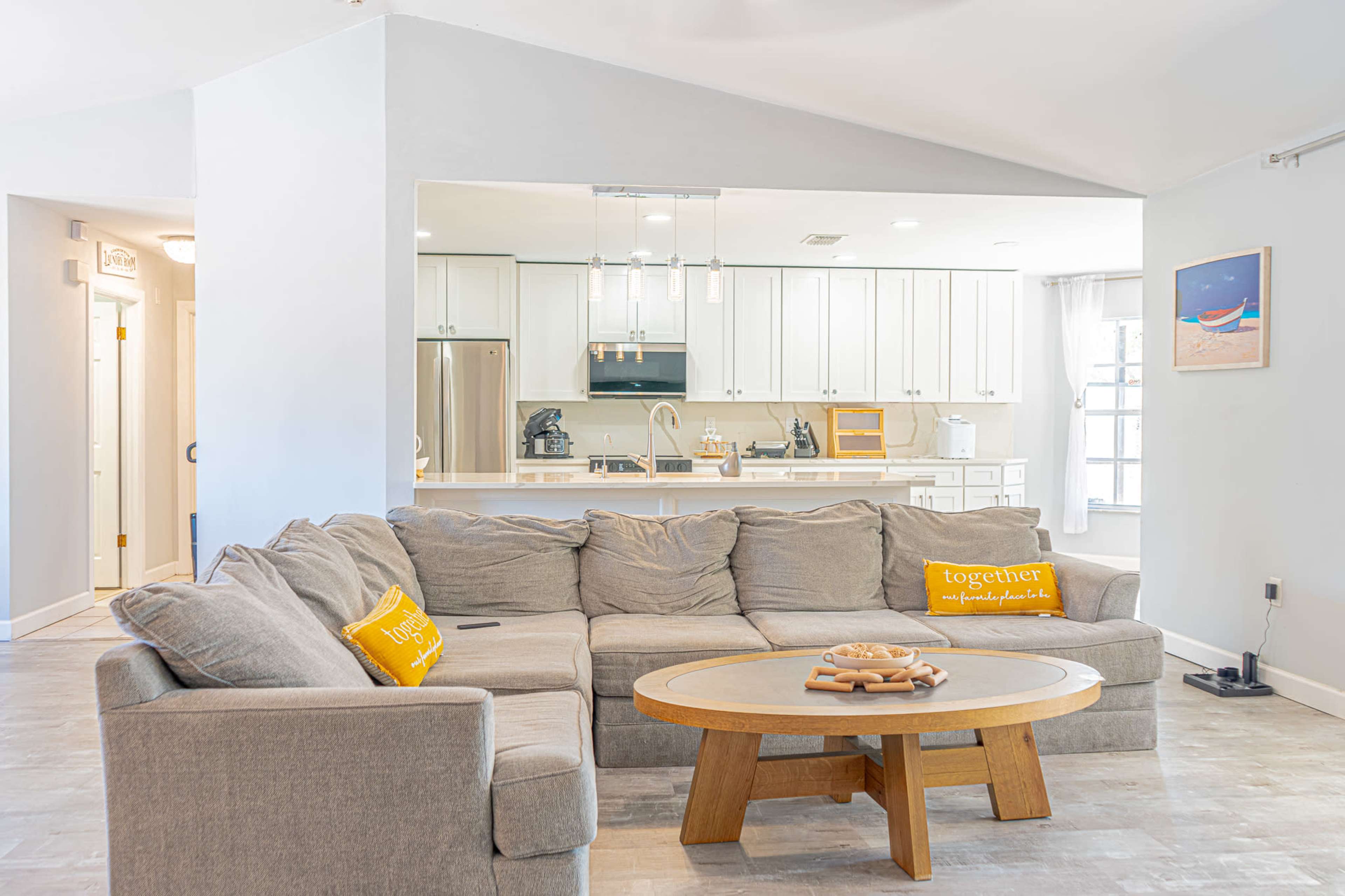 A sectional gray couch with yellow throw pillows faces a wooden coffee table in a bright and airy living space, which opens to a modern kitchen.