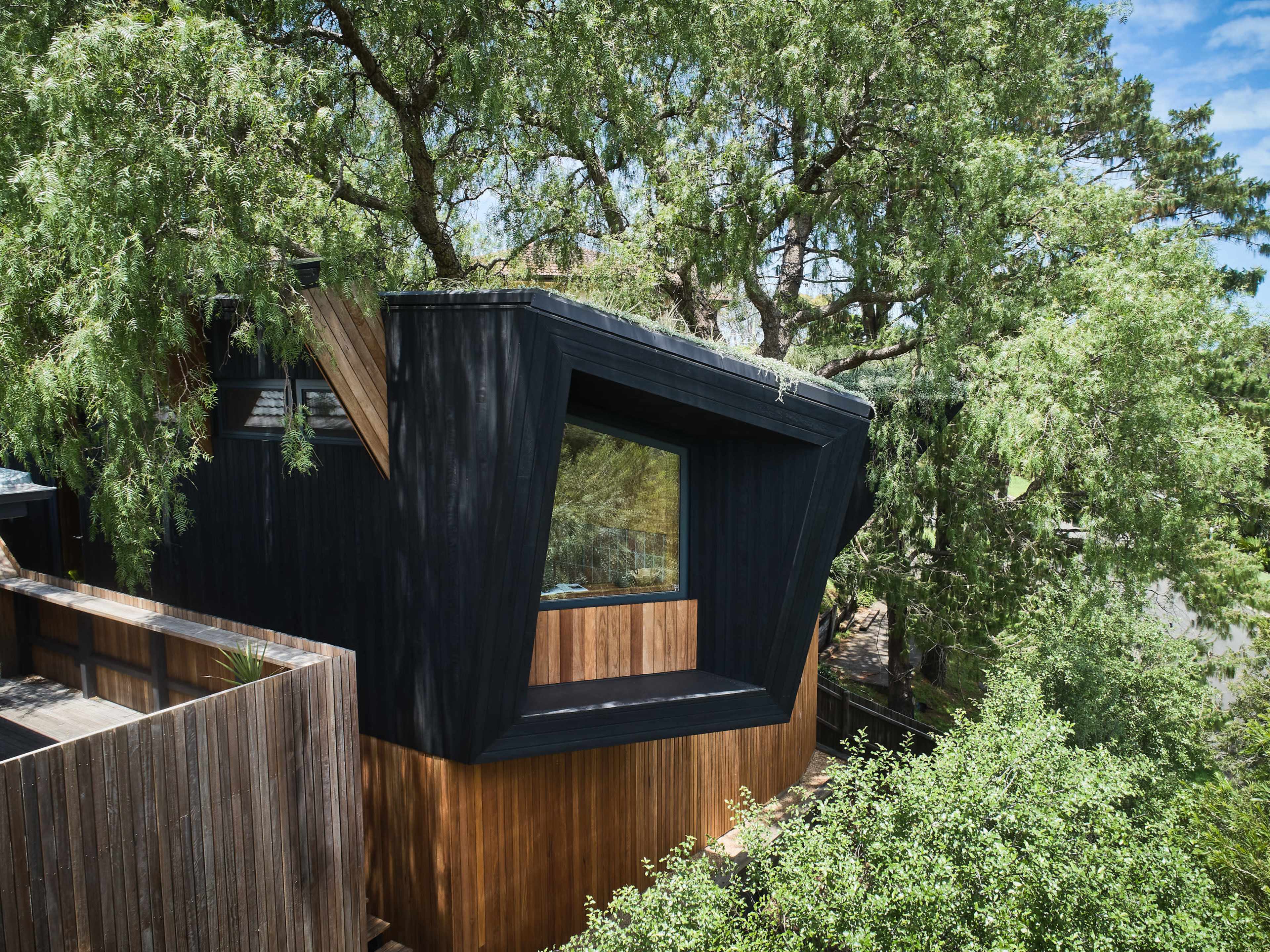 The image shows a modern black and wooden house partially surrounded by green trees.