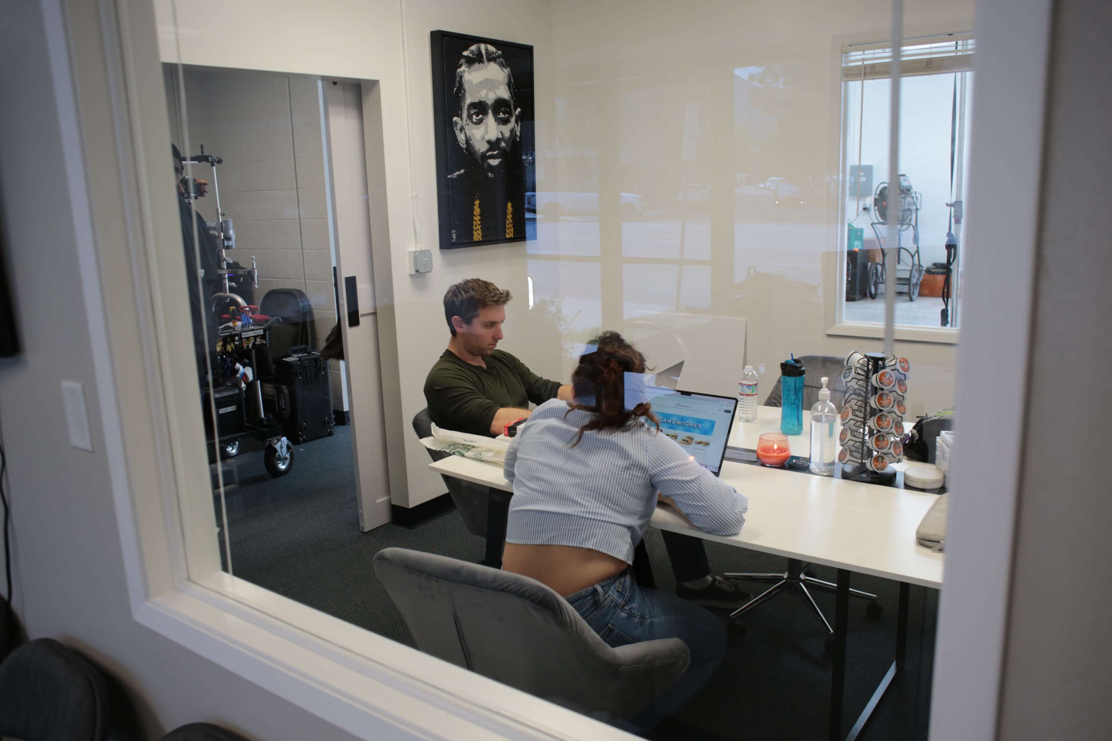 Two people work at a table in an office, with one person focused on a laptop and the other looking at a notebook, while a large portrait hangs on the wall.