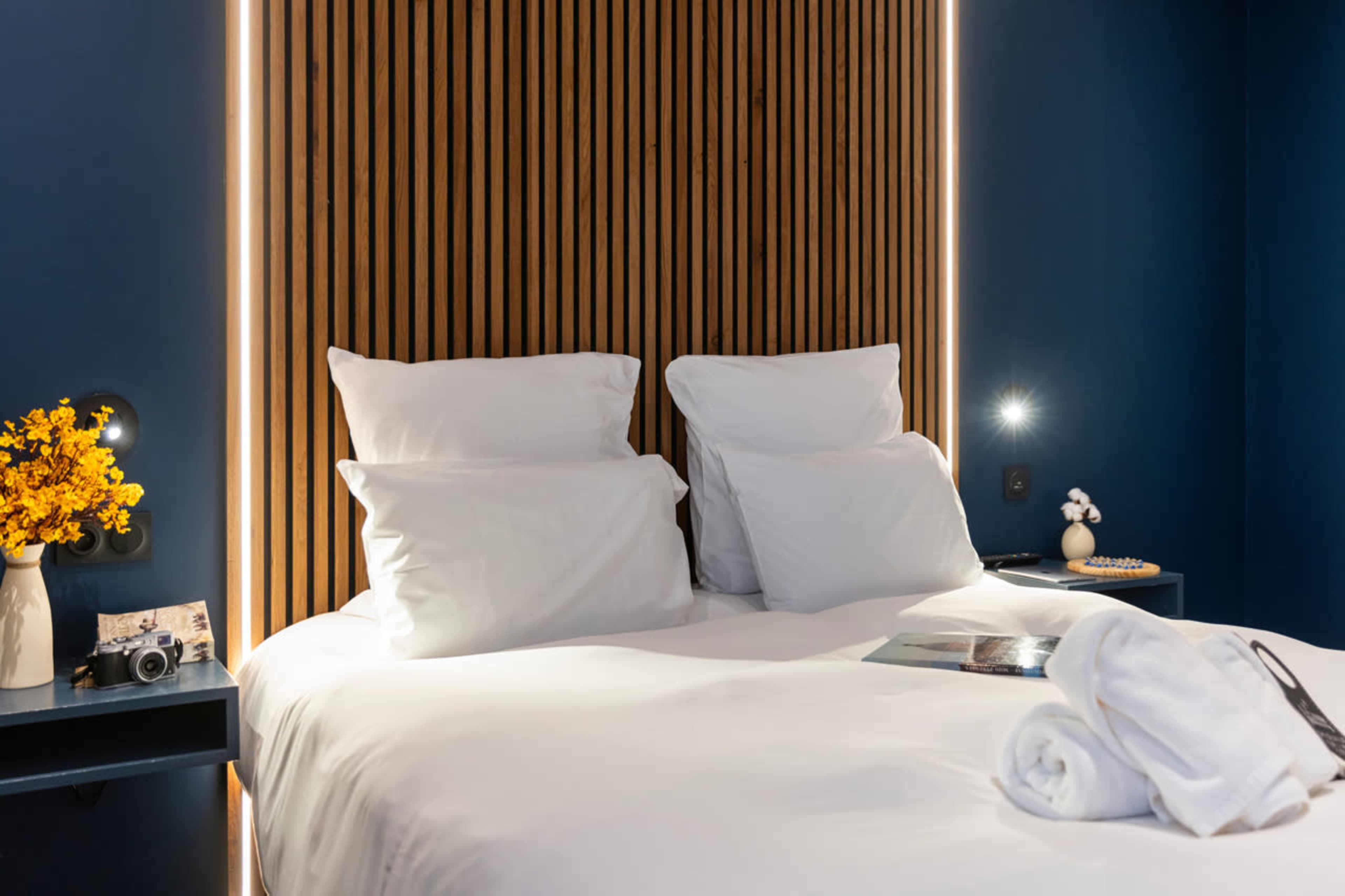 The image shows a modern hotel room with a wooden accent wall behind a neatly made bed featuring white pillows and a bedside table.
