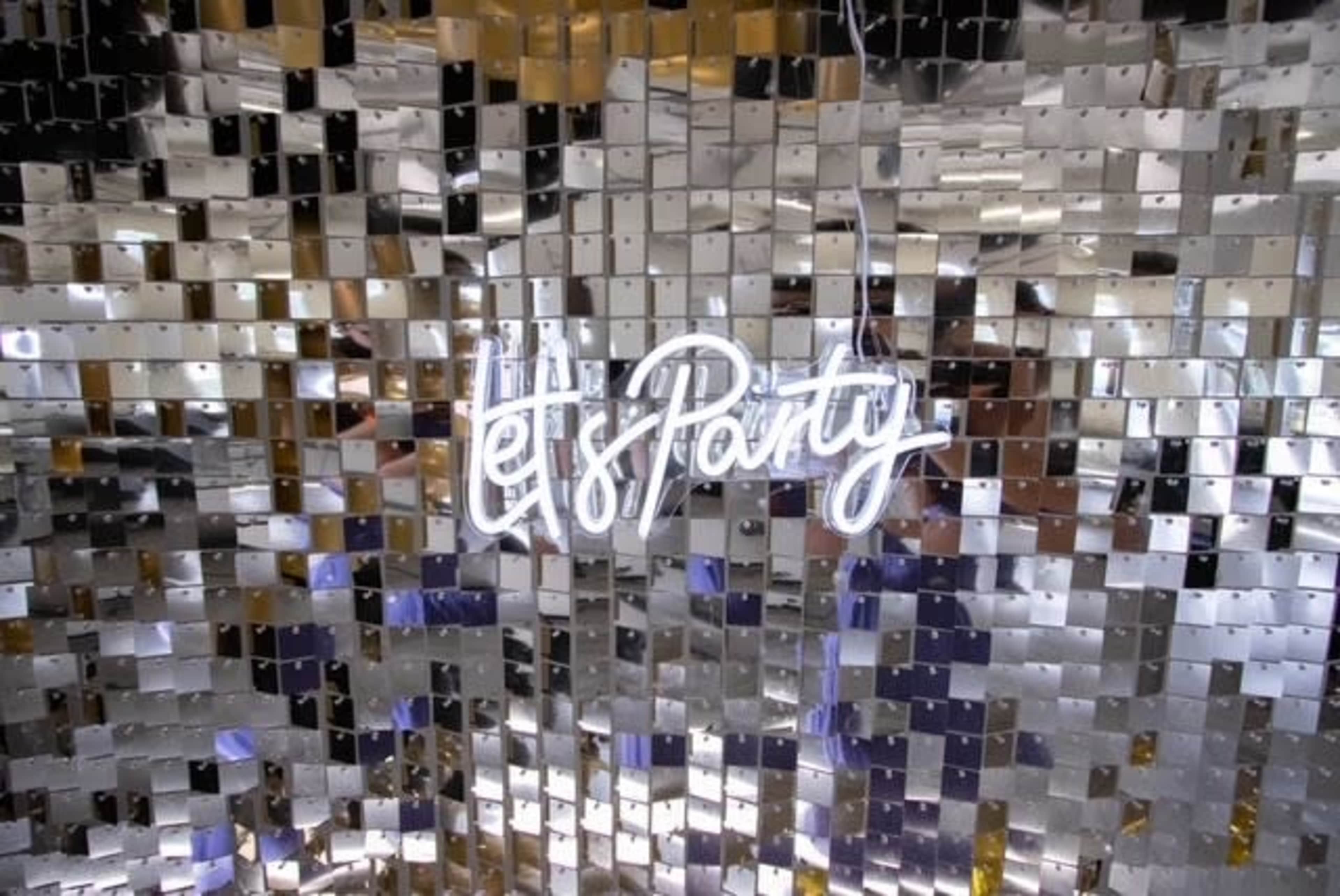 A neon sign that reads "Let's Party" is displayed against a reflective, metallic backdrop made of small square pieces.