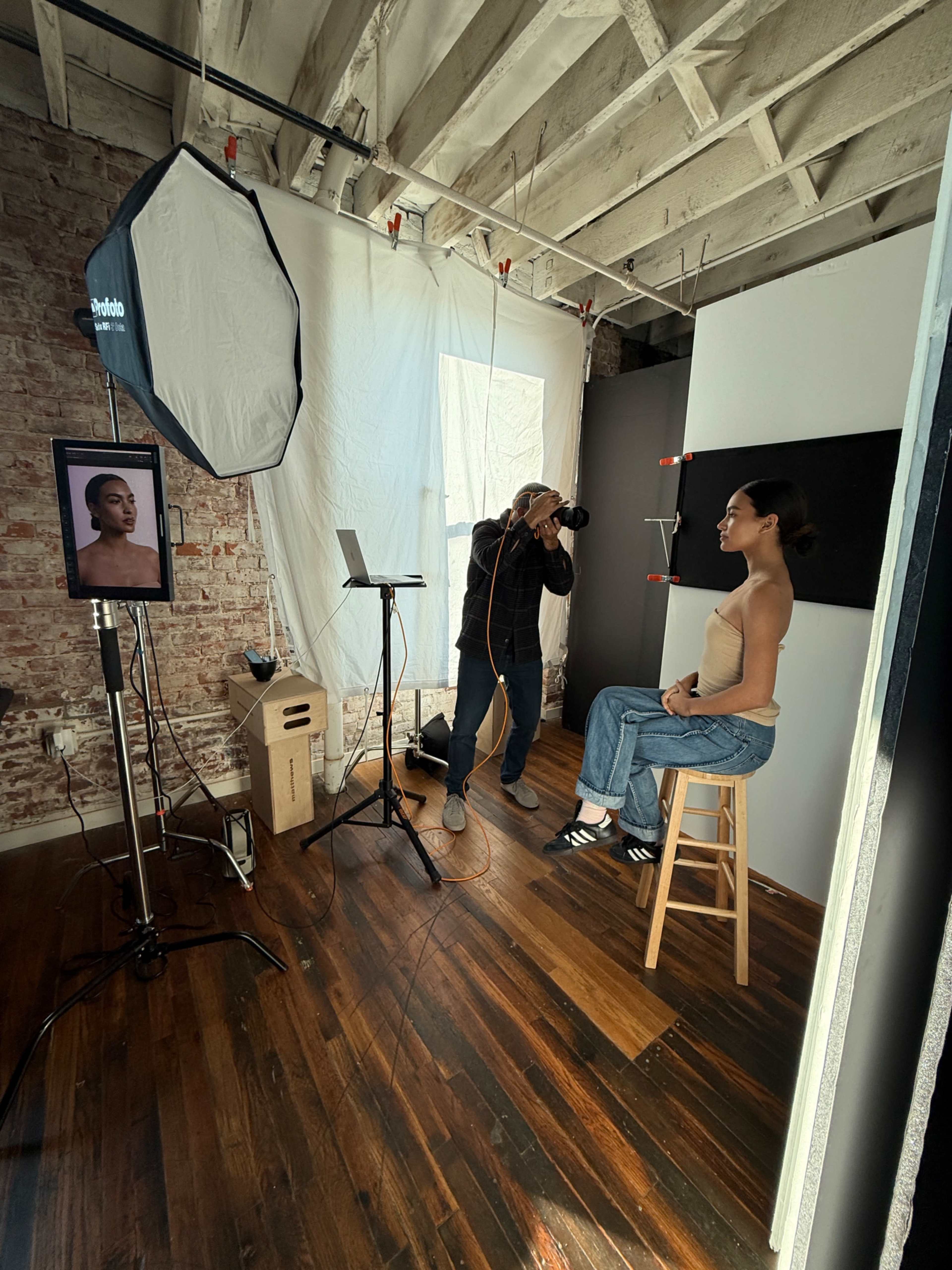 A photographer captures an image of a model seated on a stool in a well-lit studio featuring exposed brick walls and various photography equipment.