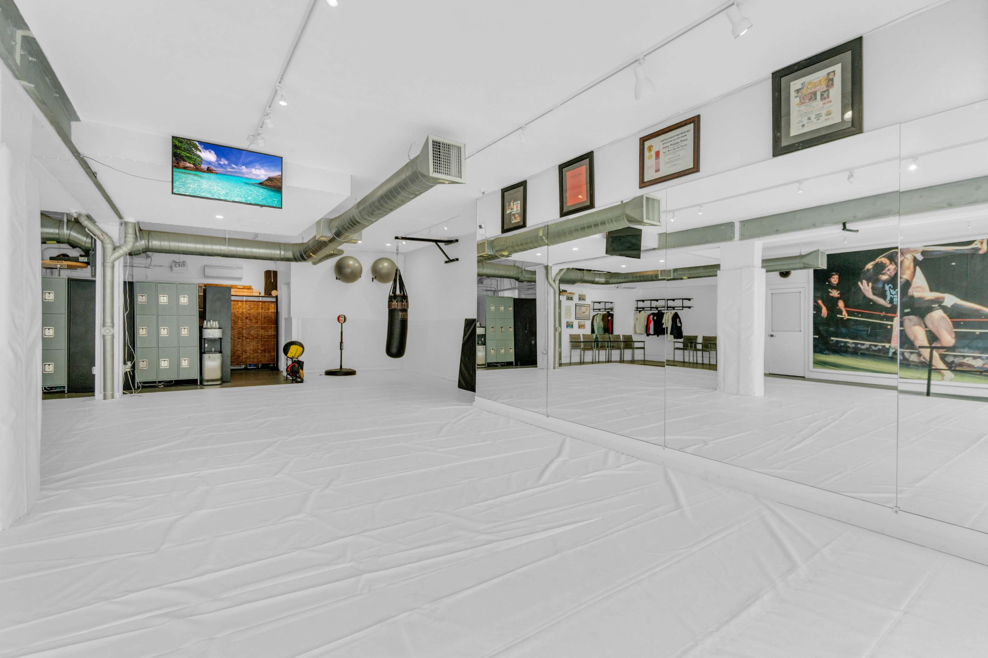 The image shows a spacious fitness studio with white flooring, mirrors on one wall, fitness equipment, and locker storage in the background.