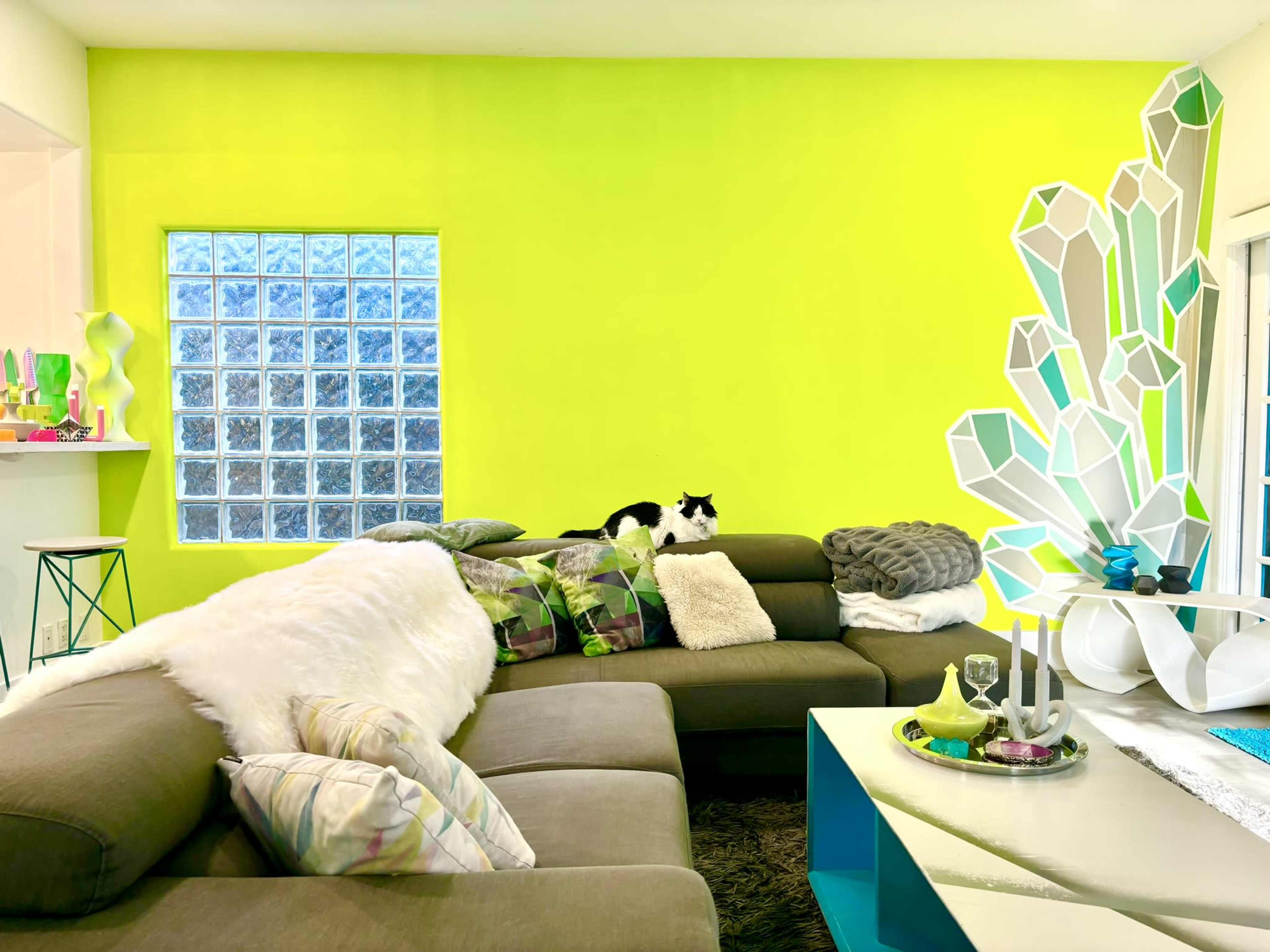 A black and white cat rests on a brown sectional sofa in a brightly colored lime green living room, which features a decorative crystal sculpture and a glass block window.