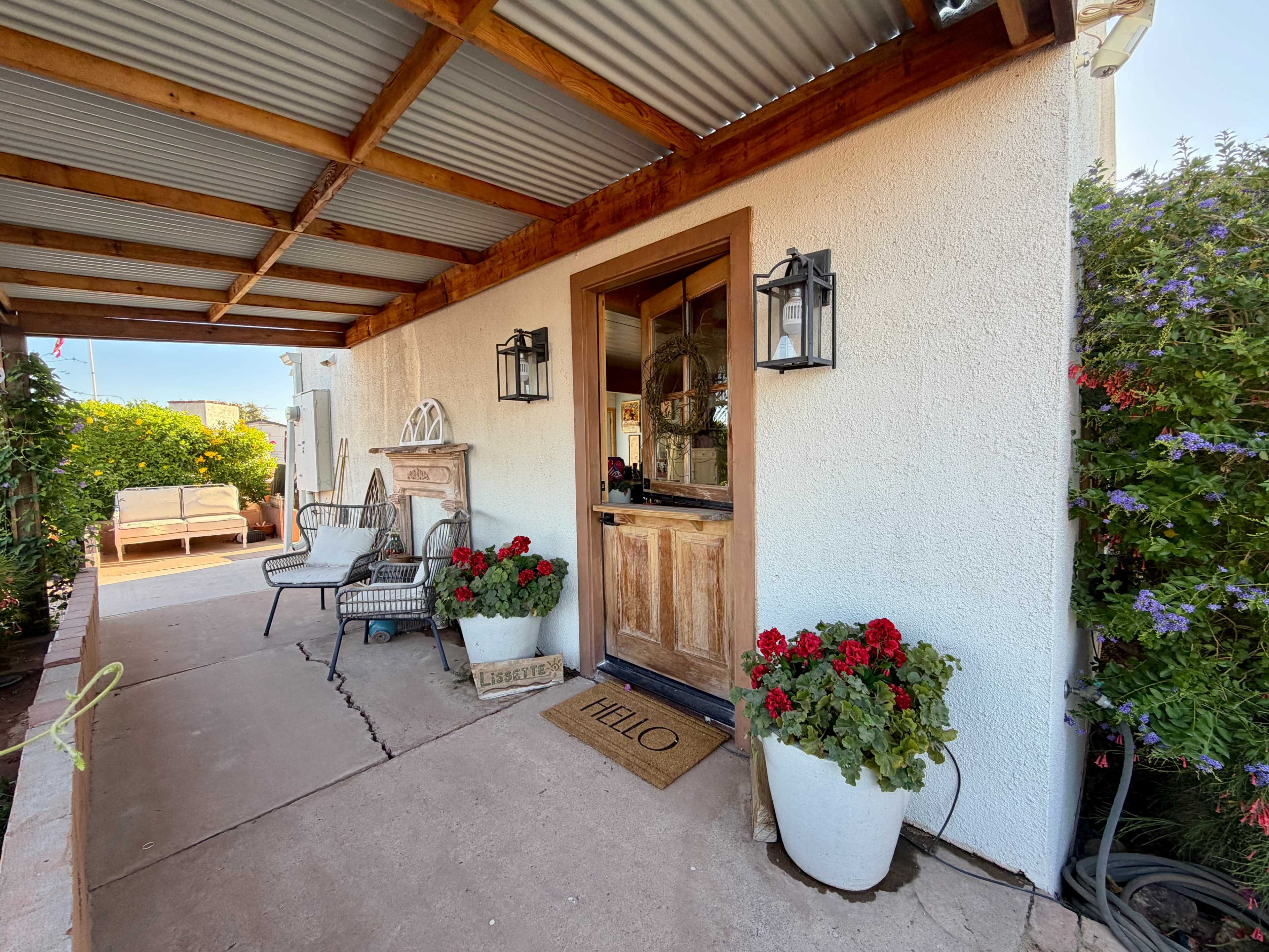 The image shows a covered porch with a wooden door framed by lanterns, flower pots, and a welcome mat.