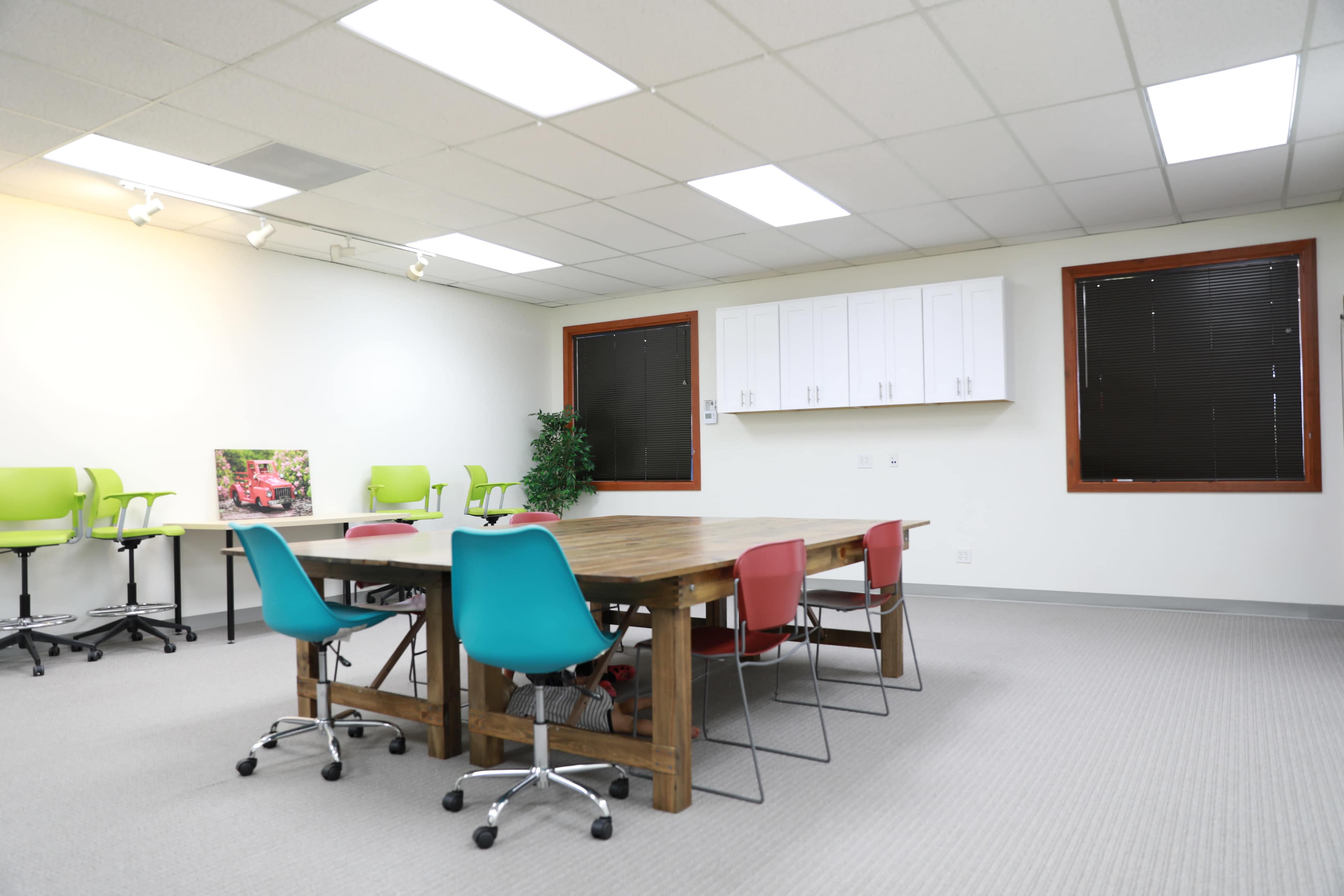 A spacious, well-lit meeting room featuring a large wooden table surrounded by colorful chairs and a few plants.