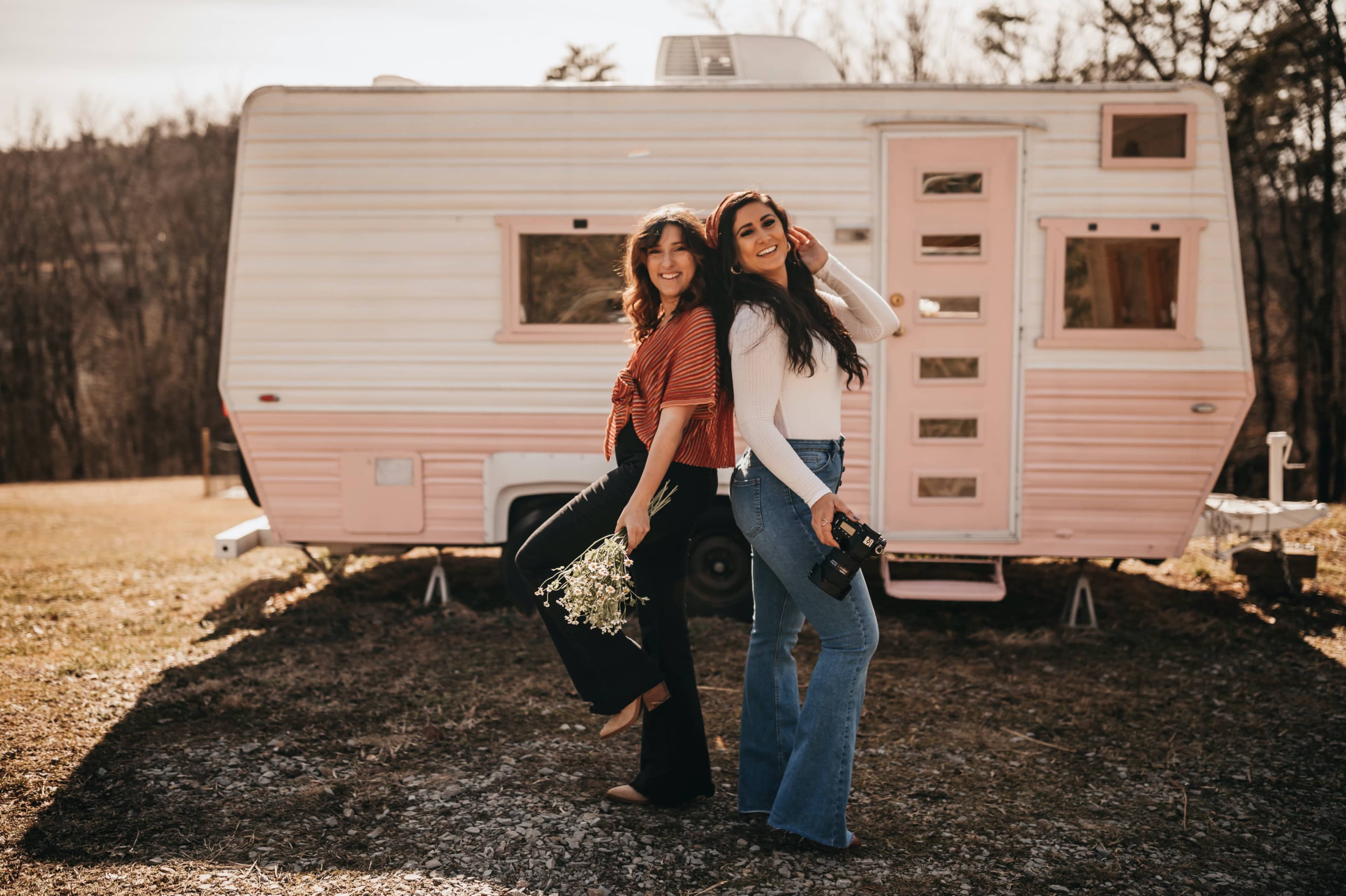 Two women pose playfully in front of a vintage pink and white camper trailer, one holding flowers and the other with a camera.