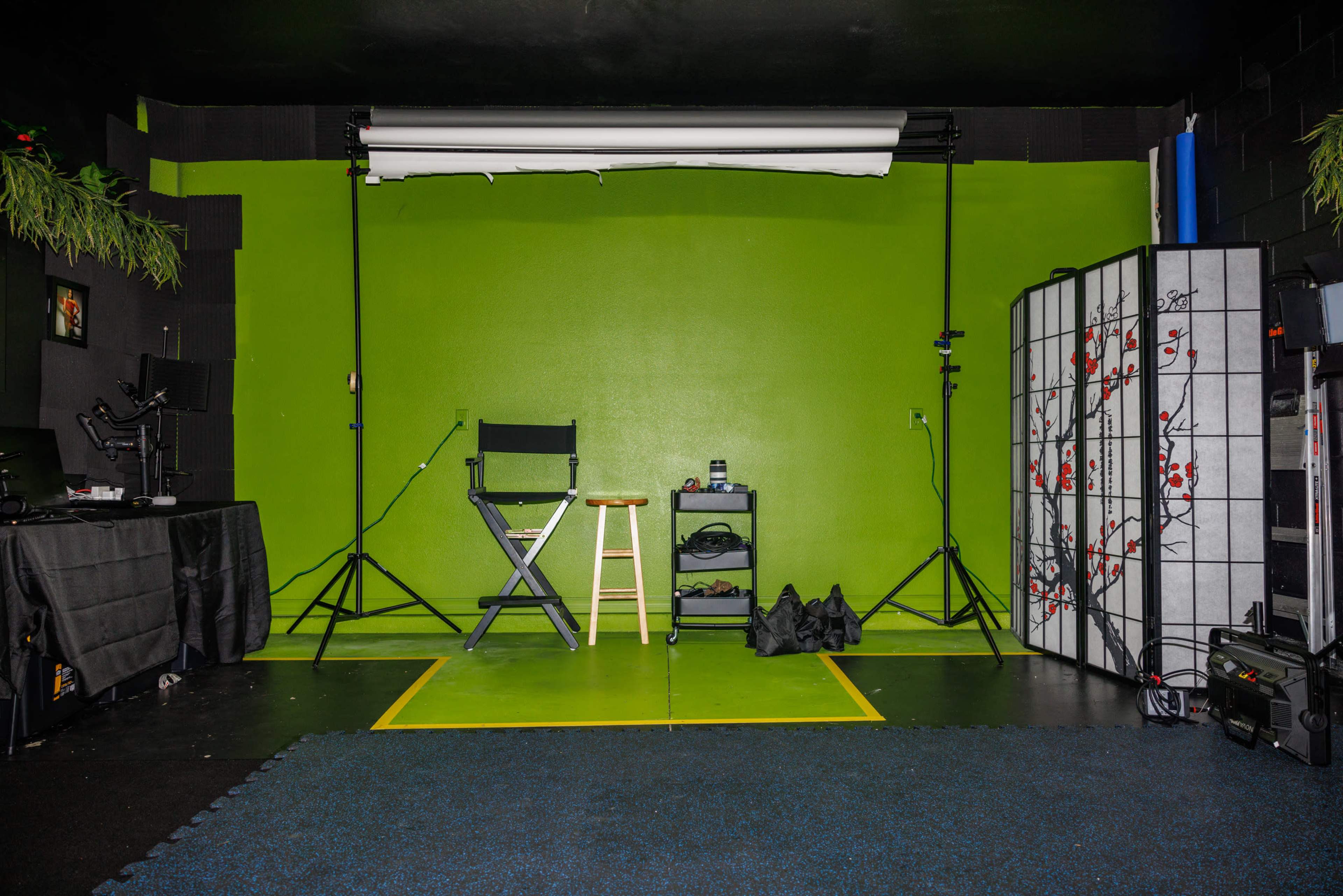 A photography studio with a green backdrop, a director's chair, a stool, lighting equipment, and a black folding screen with a floral design.