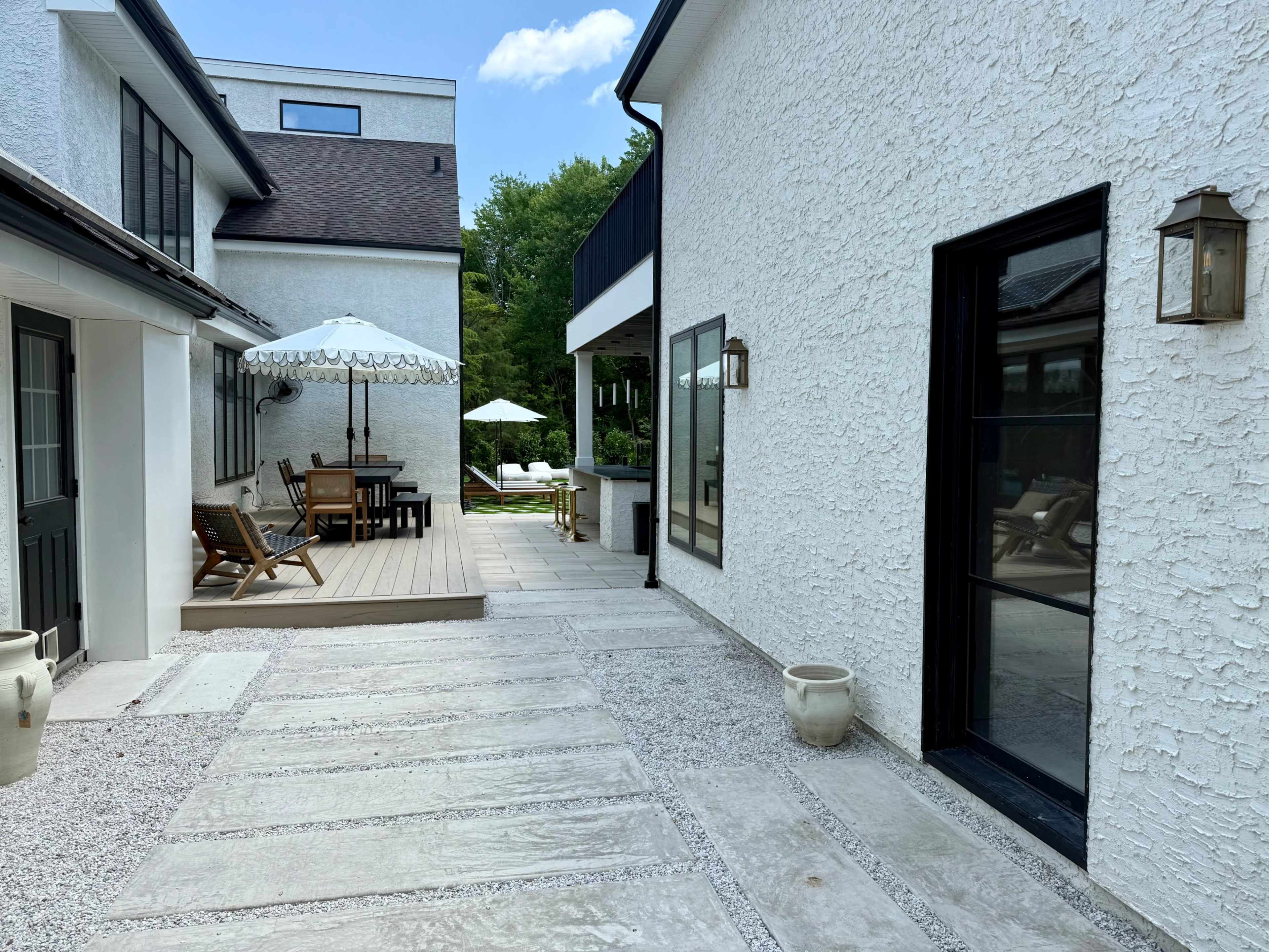 A stone pathway leads between white textured buildings to a wooden deck with outdoor seating and an umbrella.