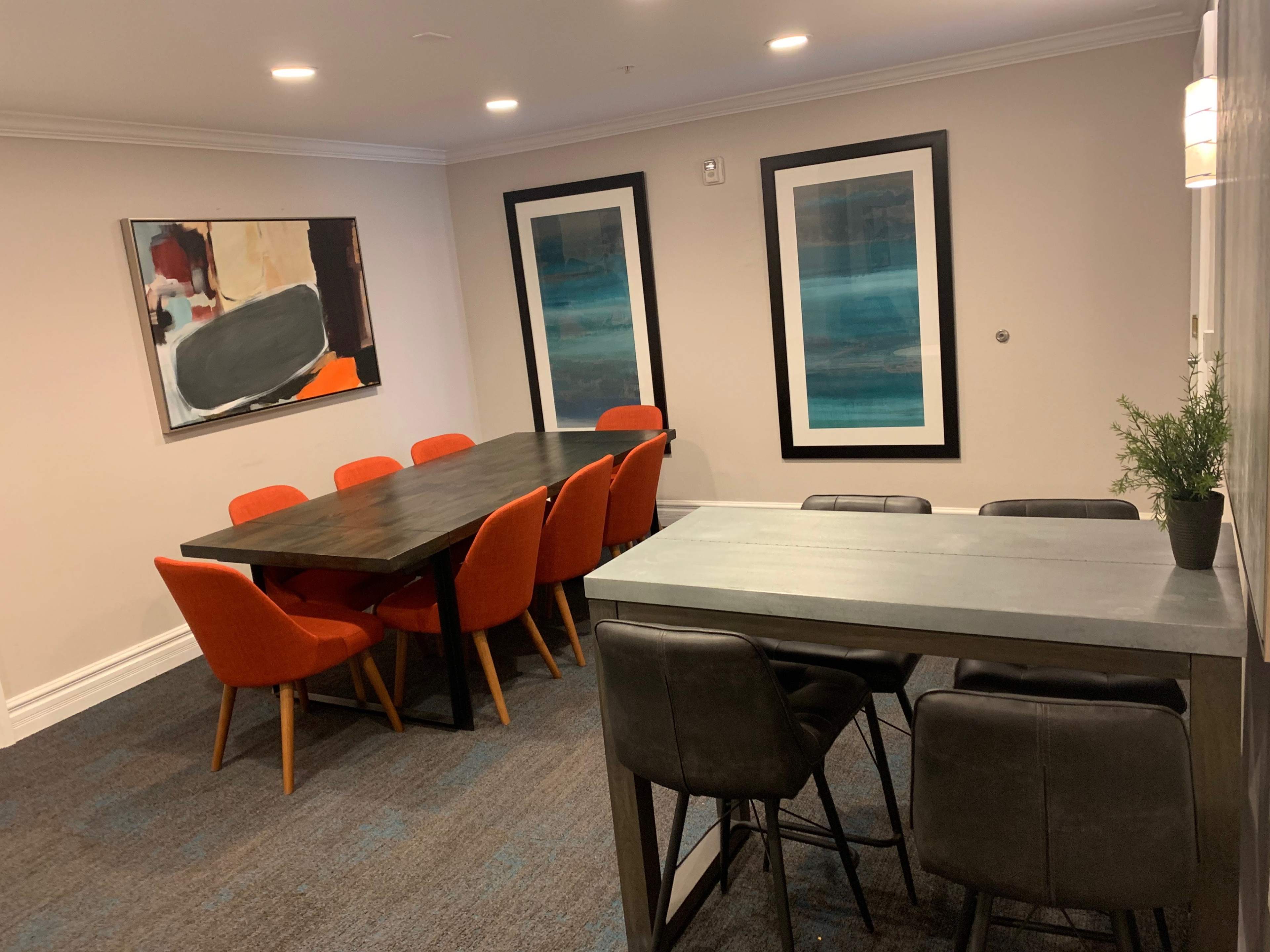 The room features a dining area with a long table surrounded by orange chairs, complemented by two framed artworks on the walls.