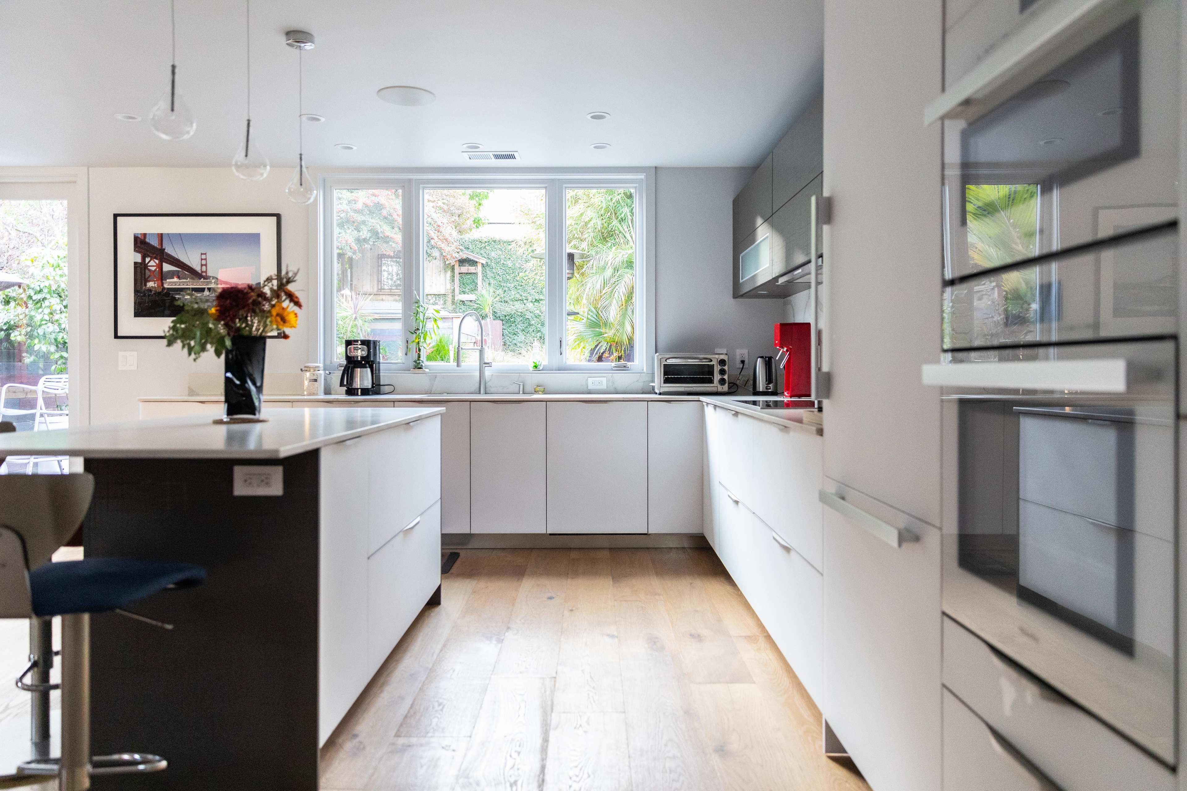 A modern kitchen features white cabinetry, a central island, and large windows overlooking a garden.
