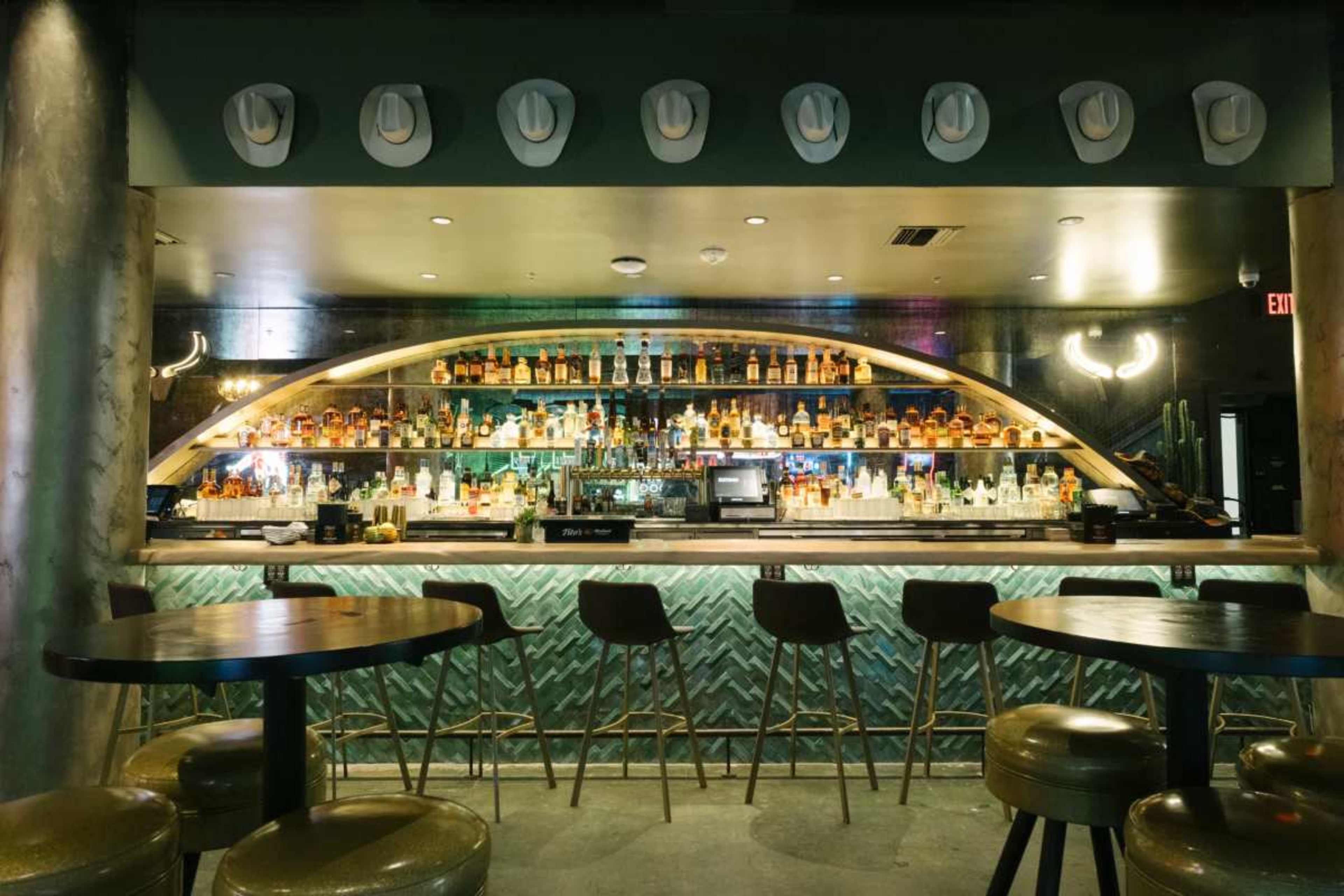 A well-stocked bar with colorful bottles lined on shelves, surrounded by high stools and decorated with cowboy hats on the wall.