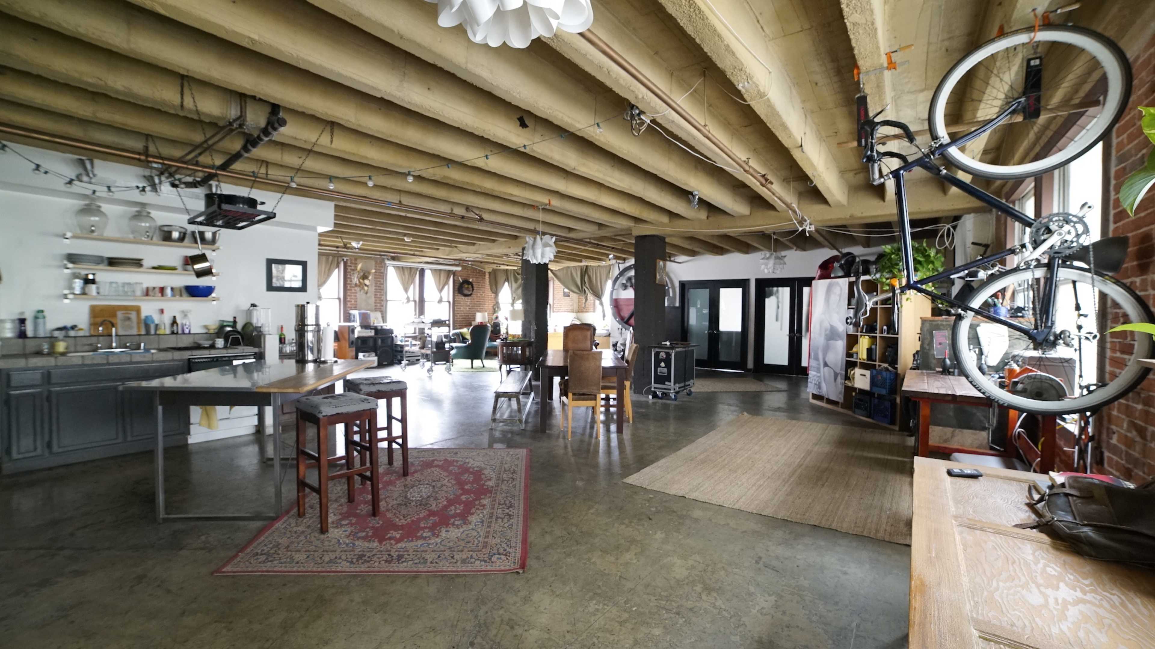 The image shows an open-concept living space with exposed wooden beams, a kitchen area, and a bicycle mounted on the wall.