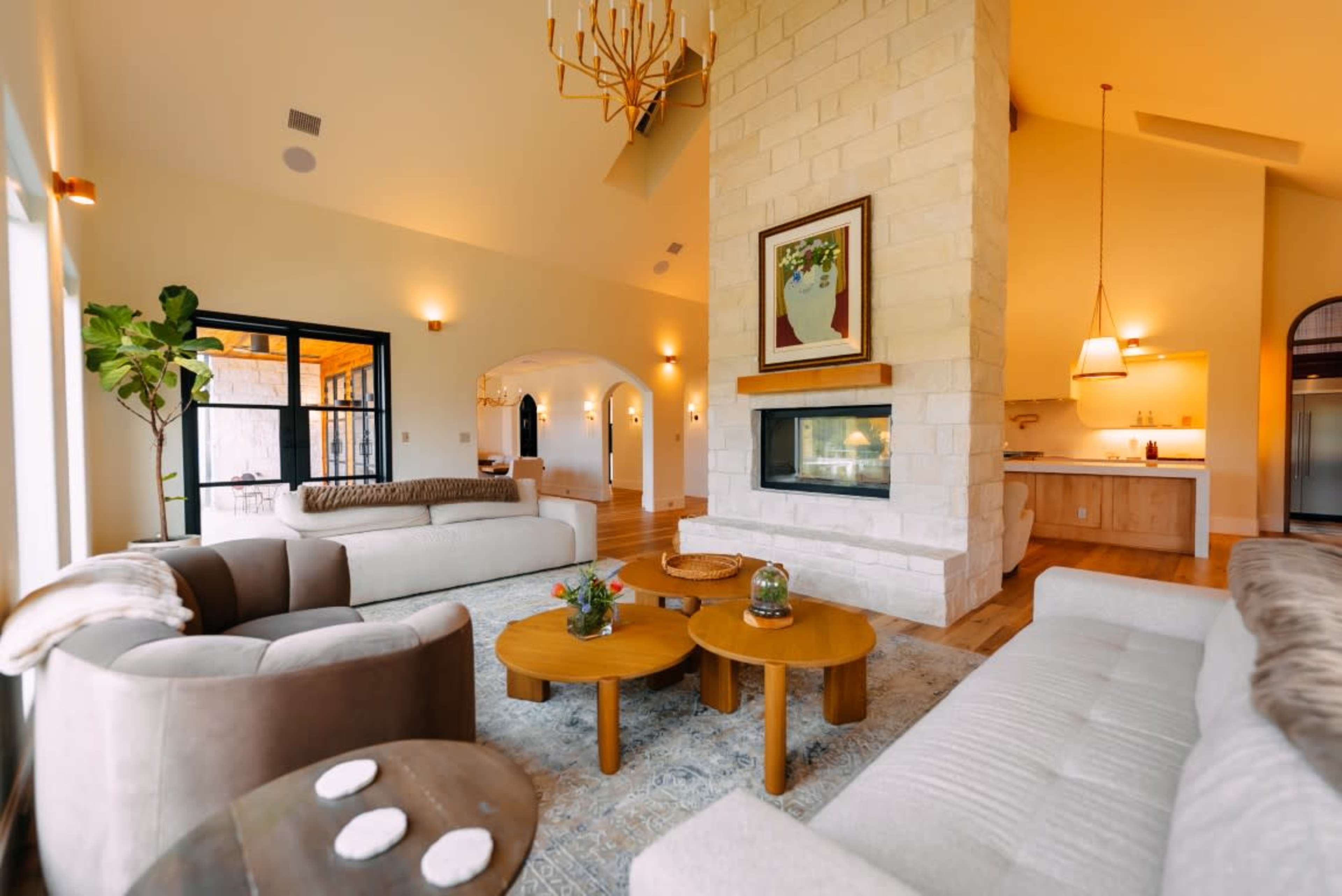 The image shows a spacious living area with a stone fireplace, comfortable seating, and large windows allowing natural light to enter the room.