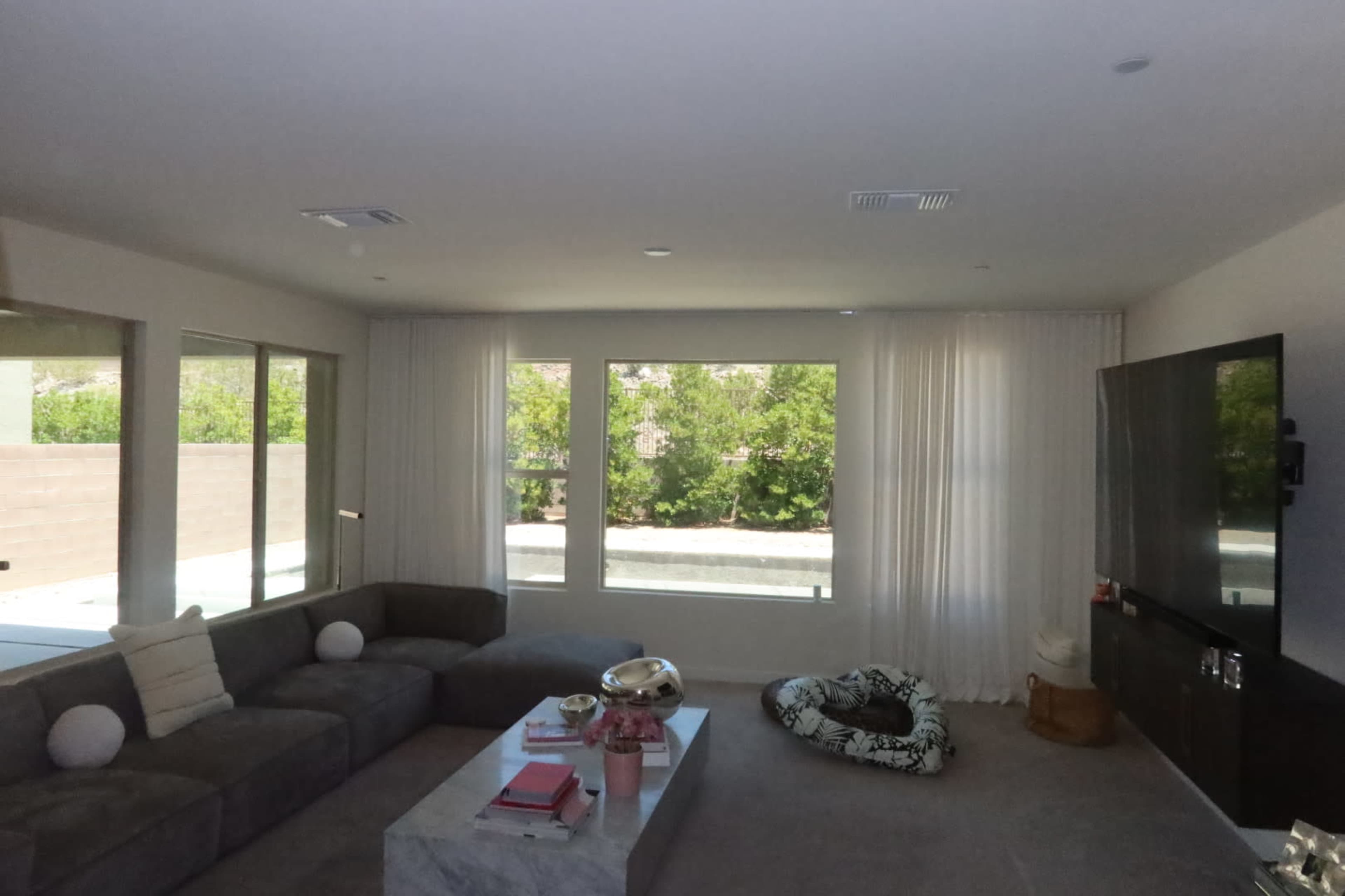 A modern living room features a large sectional sofa, a coffee table with decorative items, a window with sheer curtains, and a television unit against the wall.