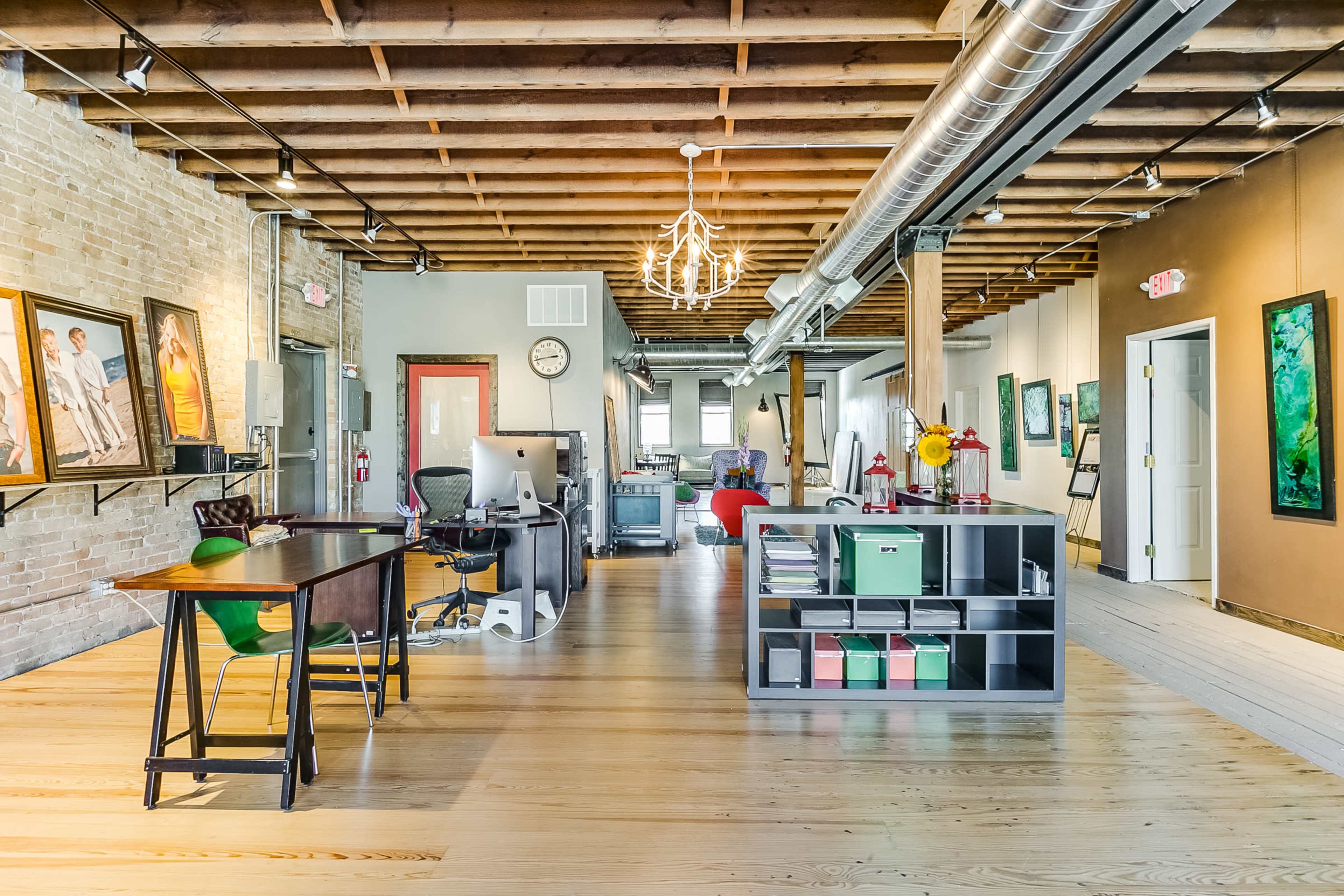 The image shows a modern office space with wooden flooring, exposed beams, and artwork displayed on the walls.