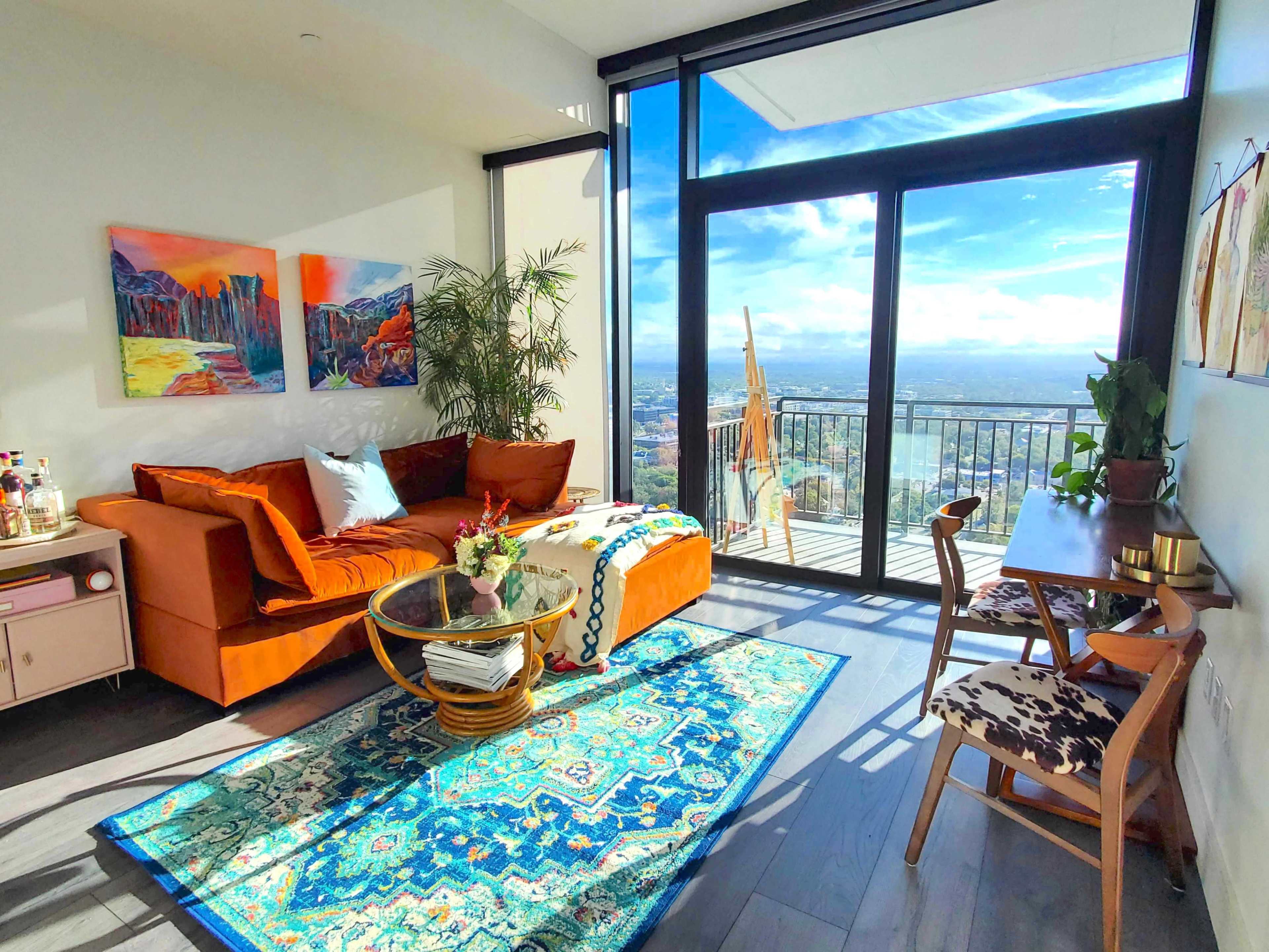A modern living room features a brown sofa, colorful artwork on the walls, a glass coffee table, and a view of the city through large windows.