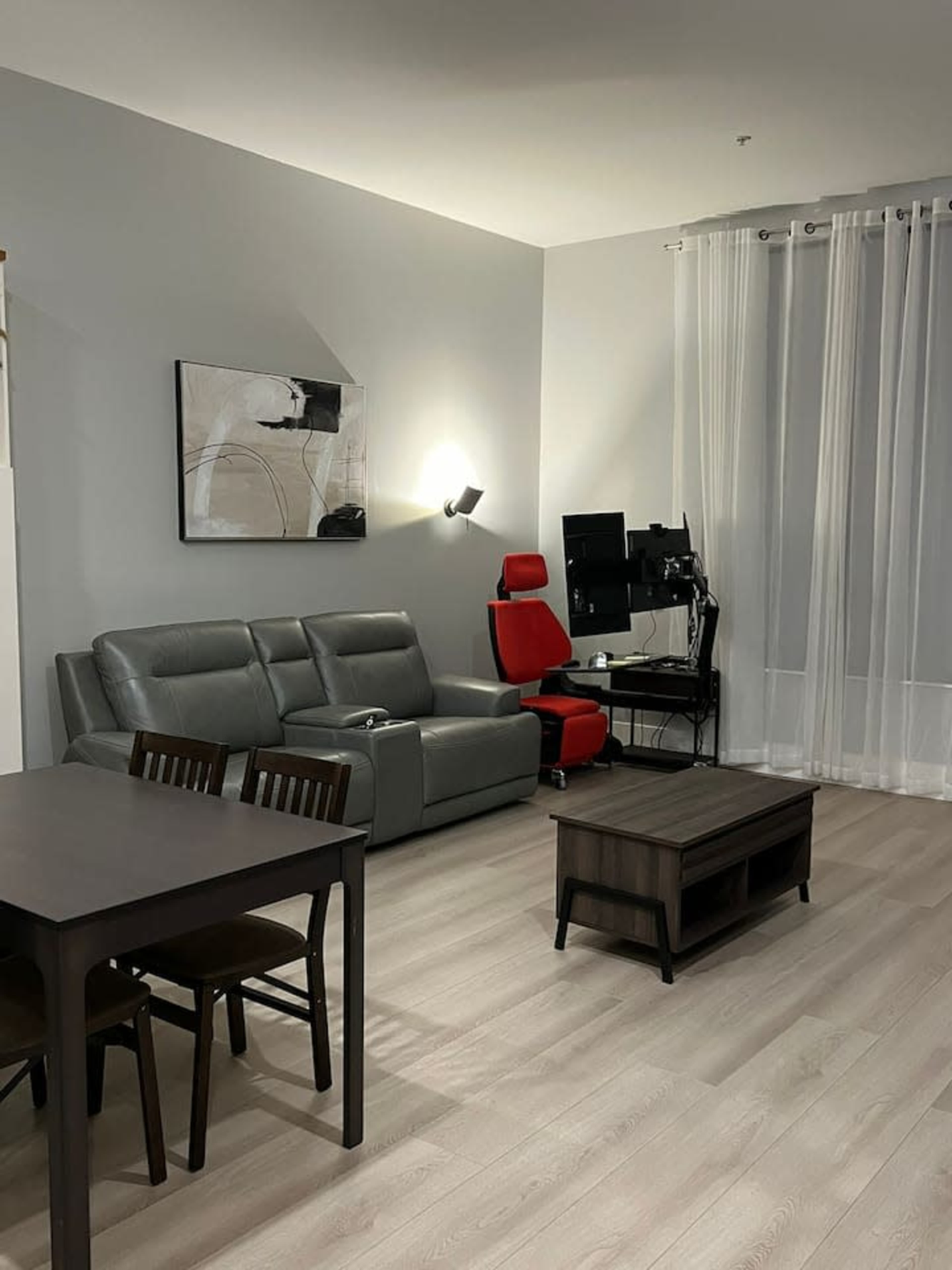 The image shows a modern living room with a grey sectional sofa, a red chair, a wooden coffee table, and a workspace featuring a computer setup, all set against light-colored walls and flooring.