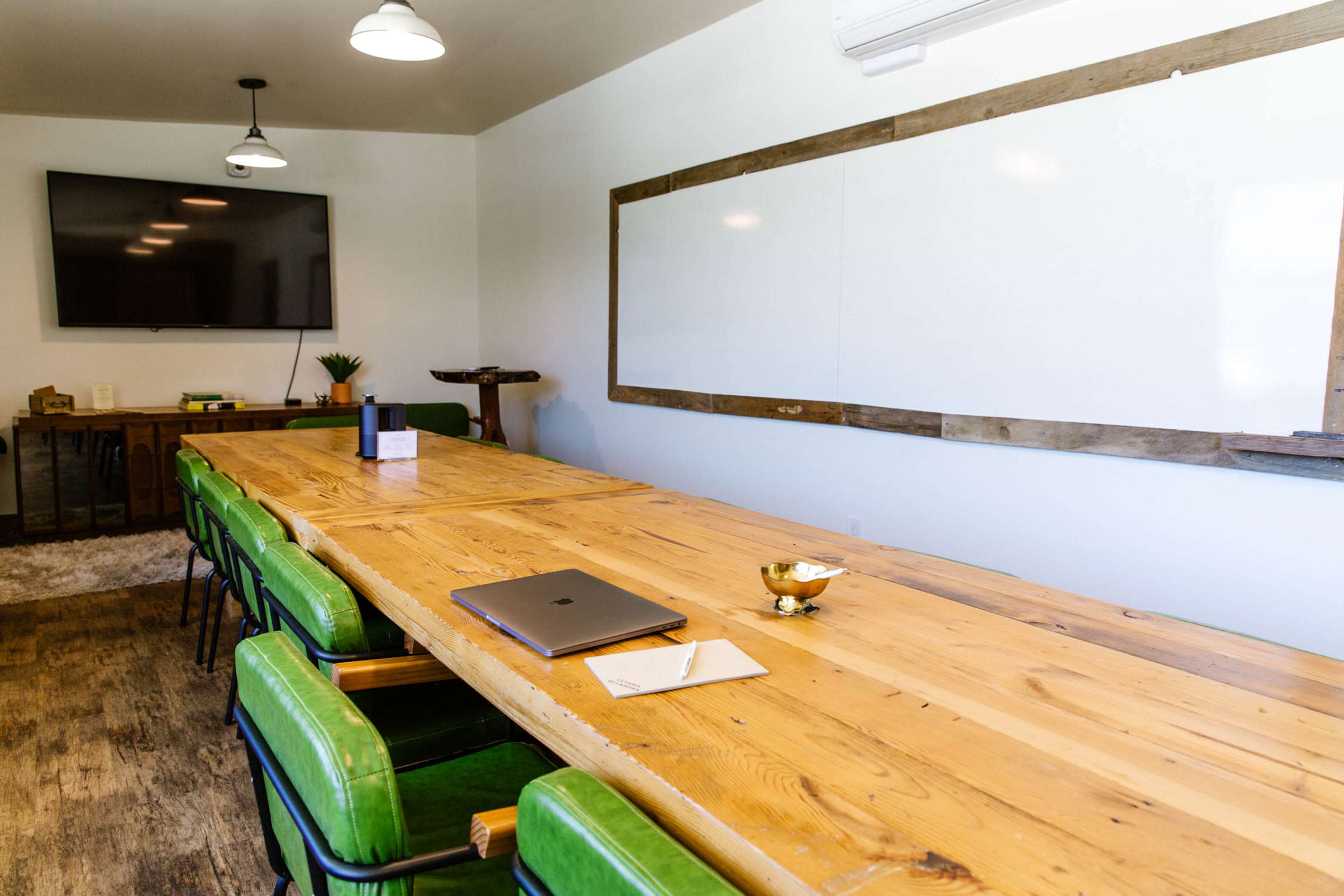 A long wooden conference table with green chairs and a laptop sits in a room featuring a large TV and a whiteboard.