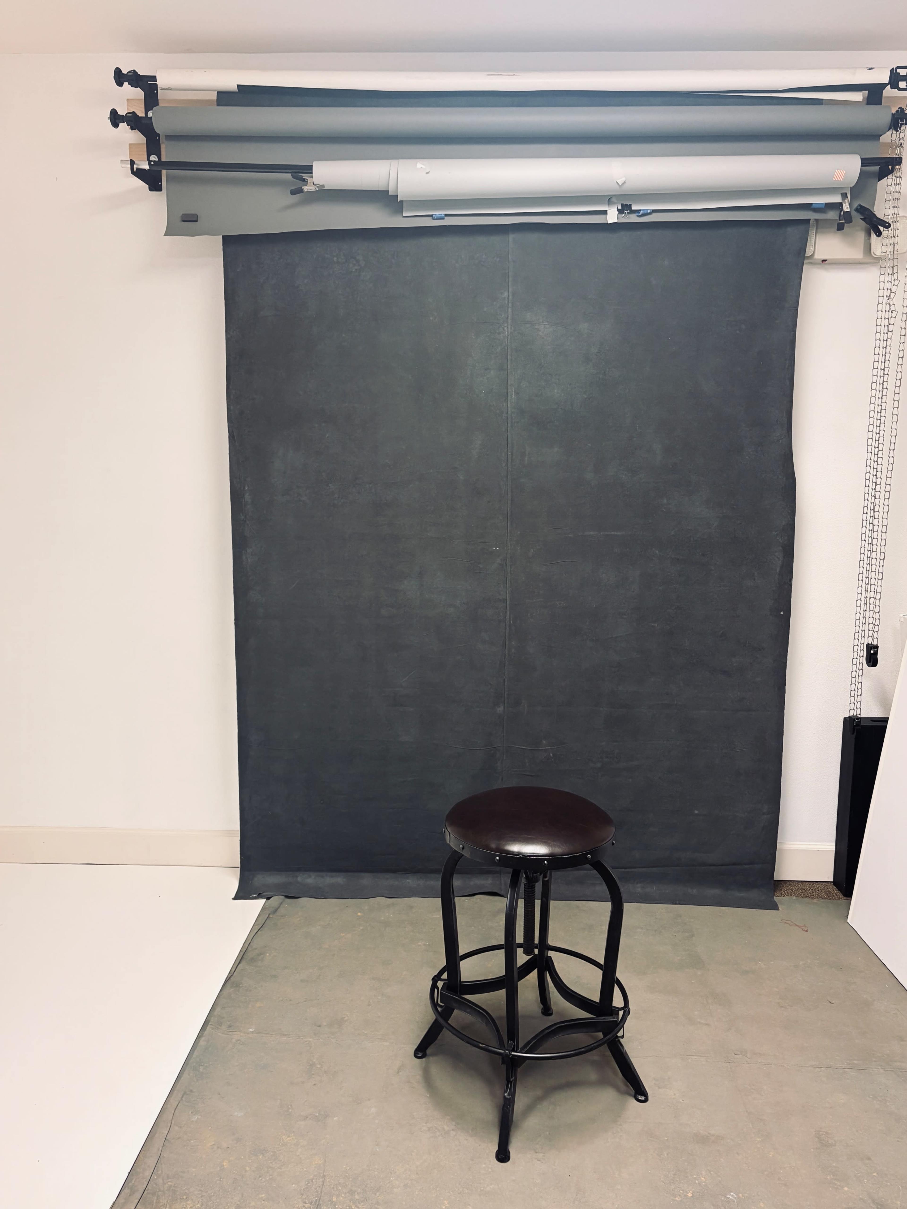 The image shows a black stool in front of a dark backdrop in a studio setting.