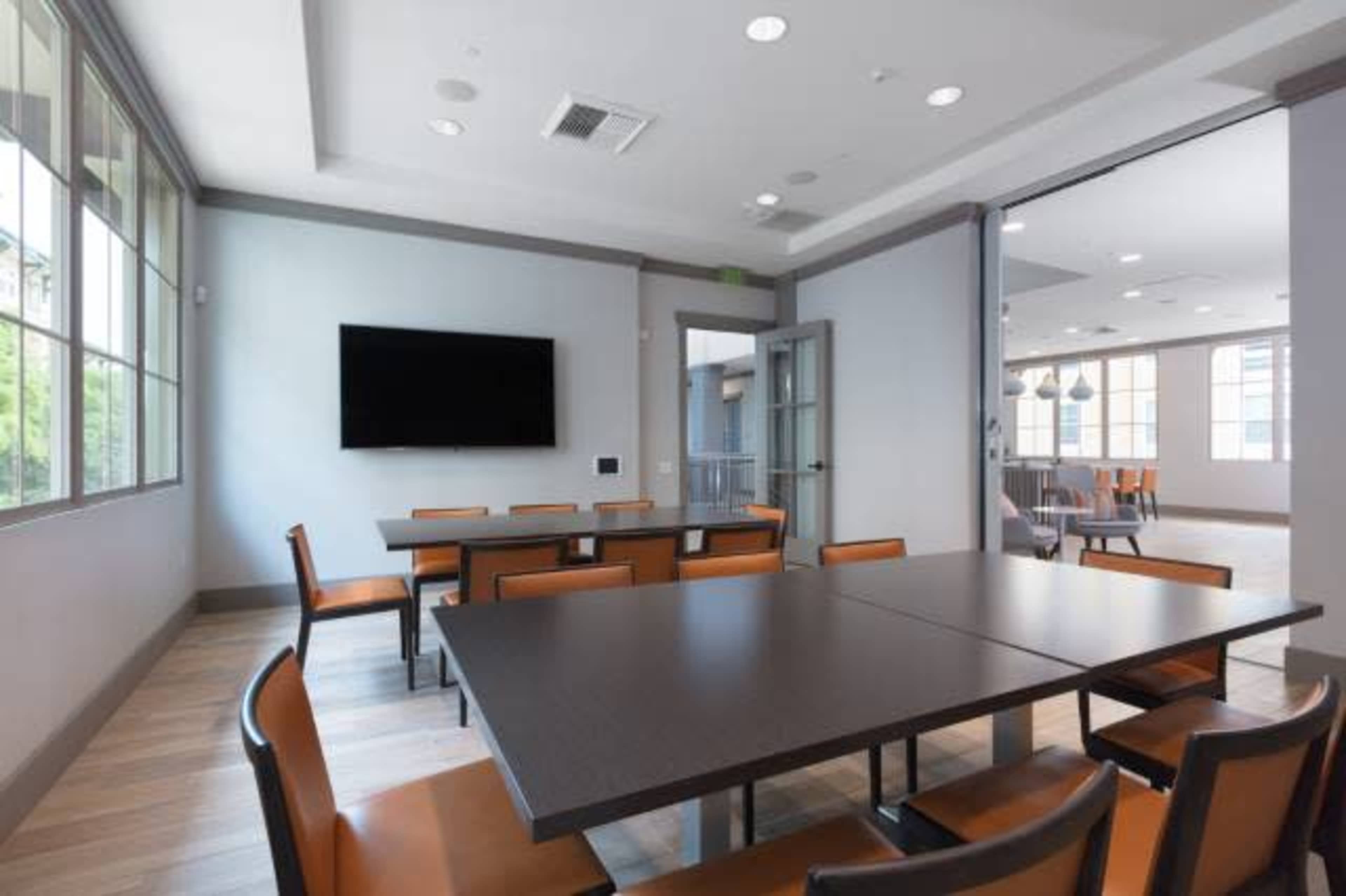 A conference room features a large table surrounded by eight brown chairs, with a television mounted on one wall and large windows providing natural light.