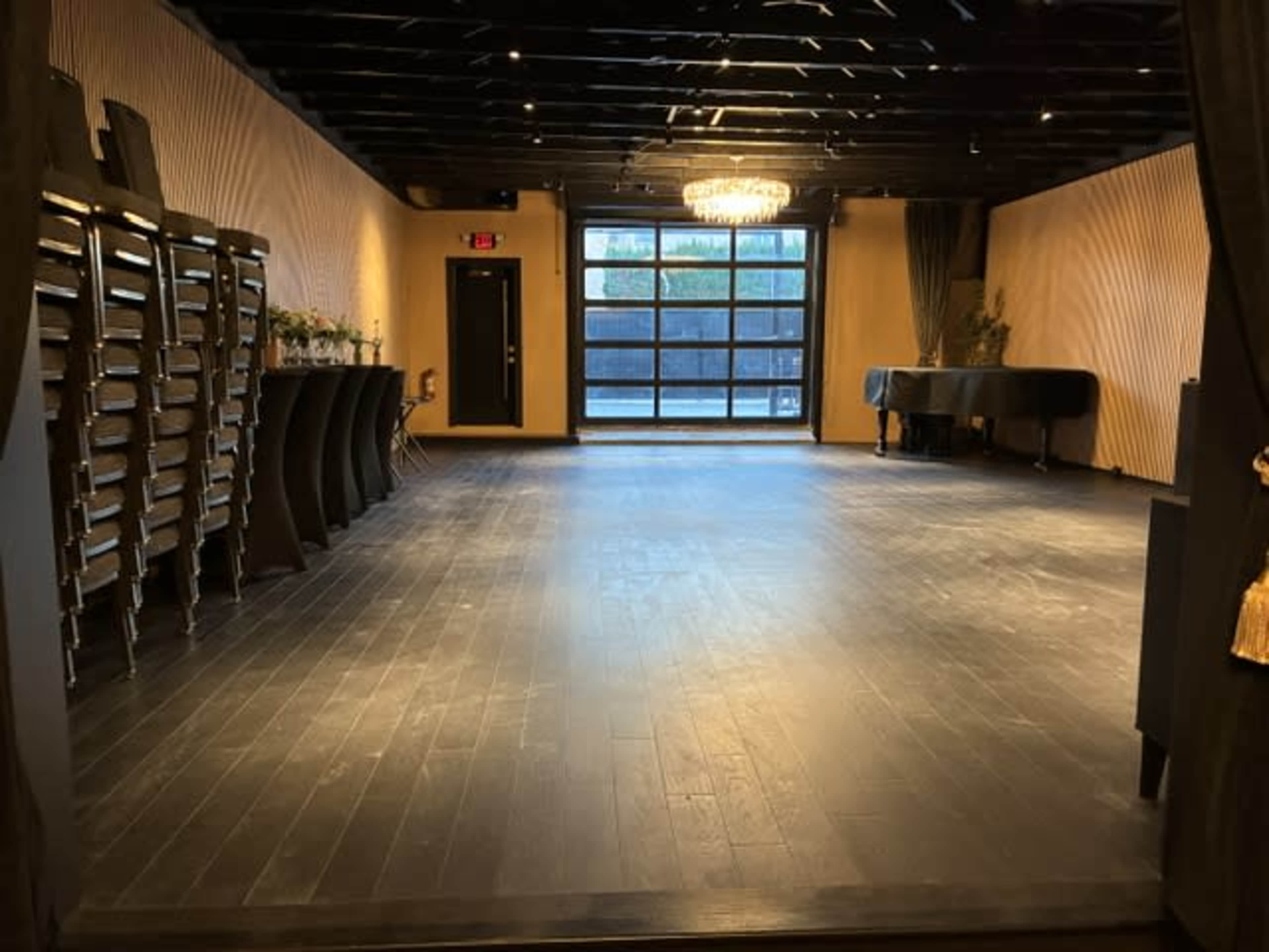 The image shows an empty room with hardwood floors, a grand piano in one corner, and stacked chairs lined against the wall, illuminated by a chandelier and natural light coming through large windows.