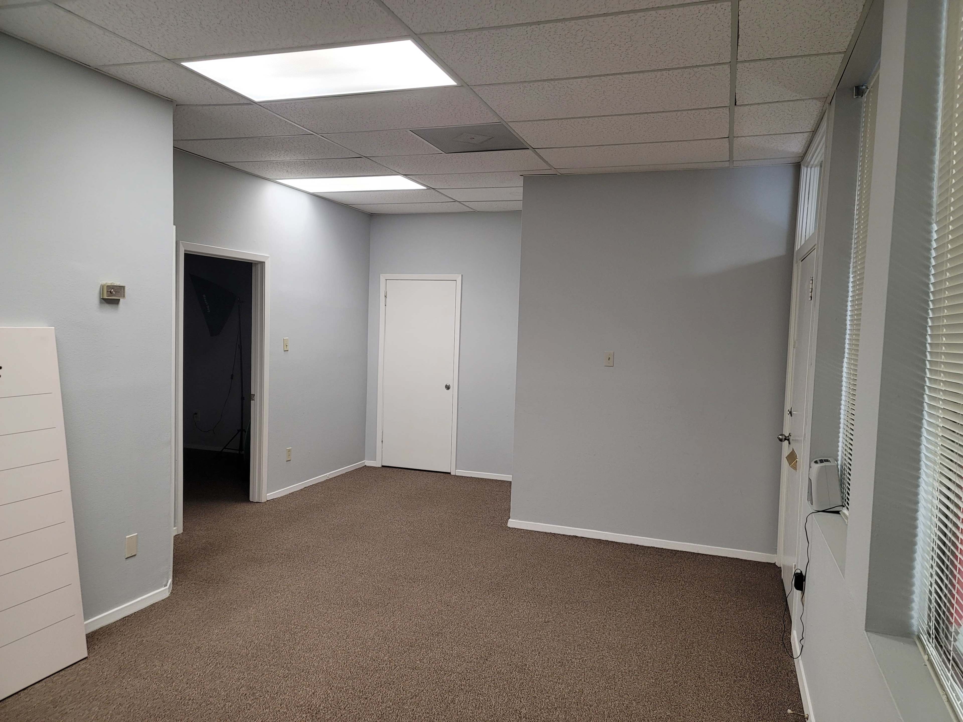 The image shows a vacant office space with light gray walls, a carpeted floor, and an interior door on one side.