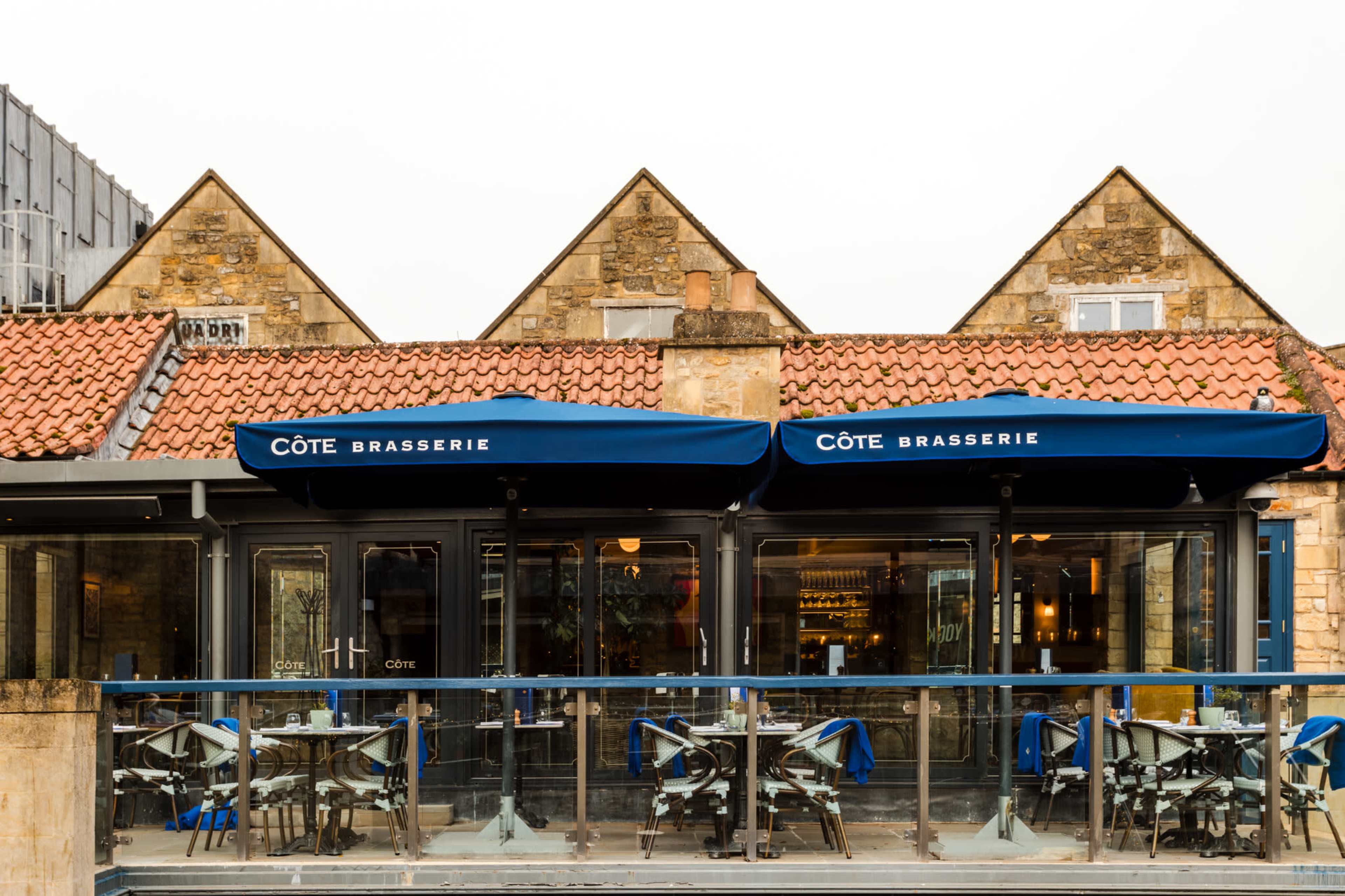 The image shows the exterior of a brasserie called Côté, featuring a glass patio with blue chairs and umbrellas, against a backdrop of stone buildings with triangular roofs.