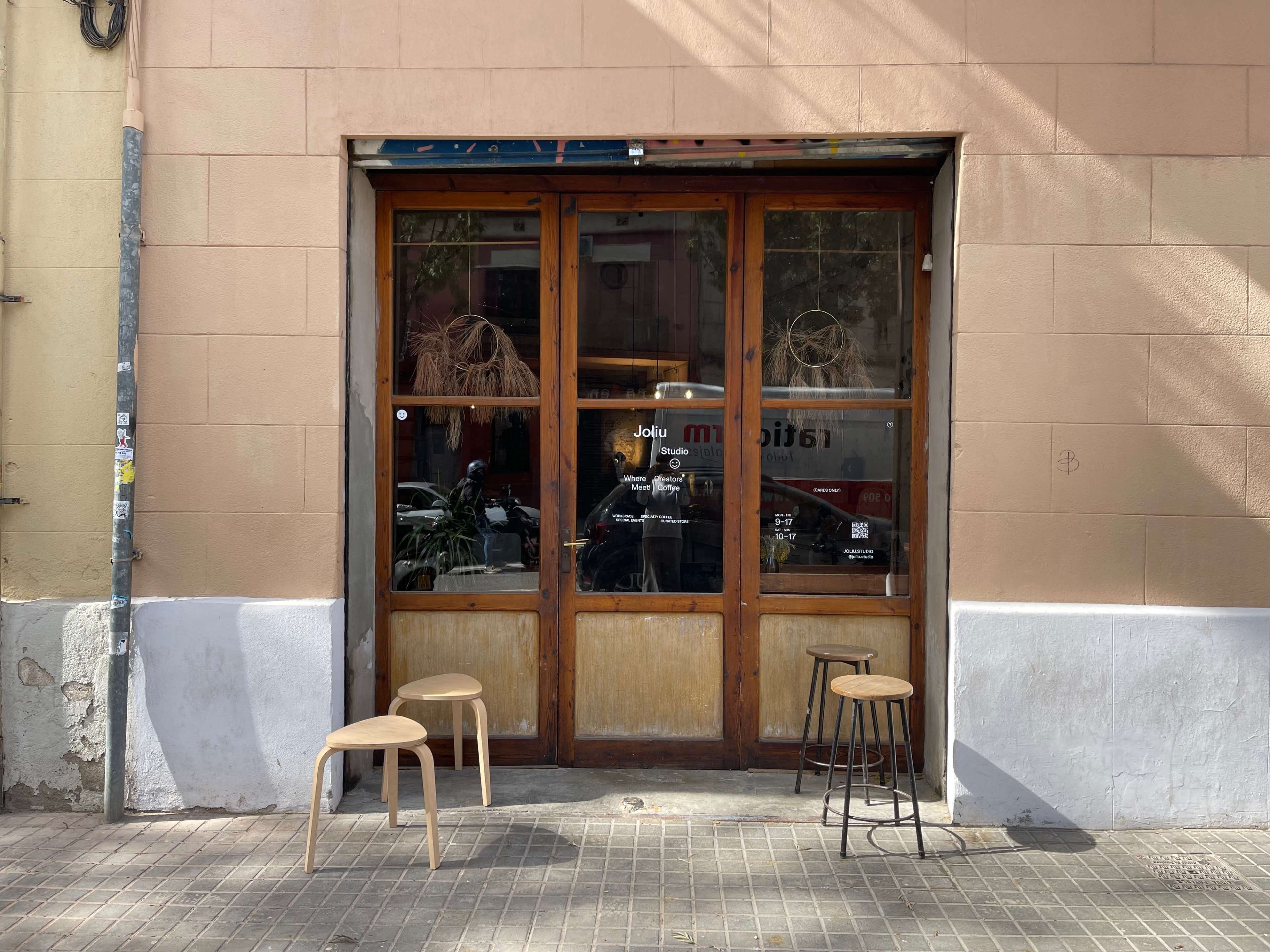 A wooden-framed storefront with large glass doors, featuring three stools on the sidewalk in front.