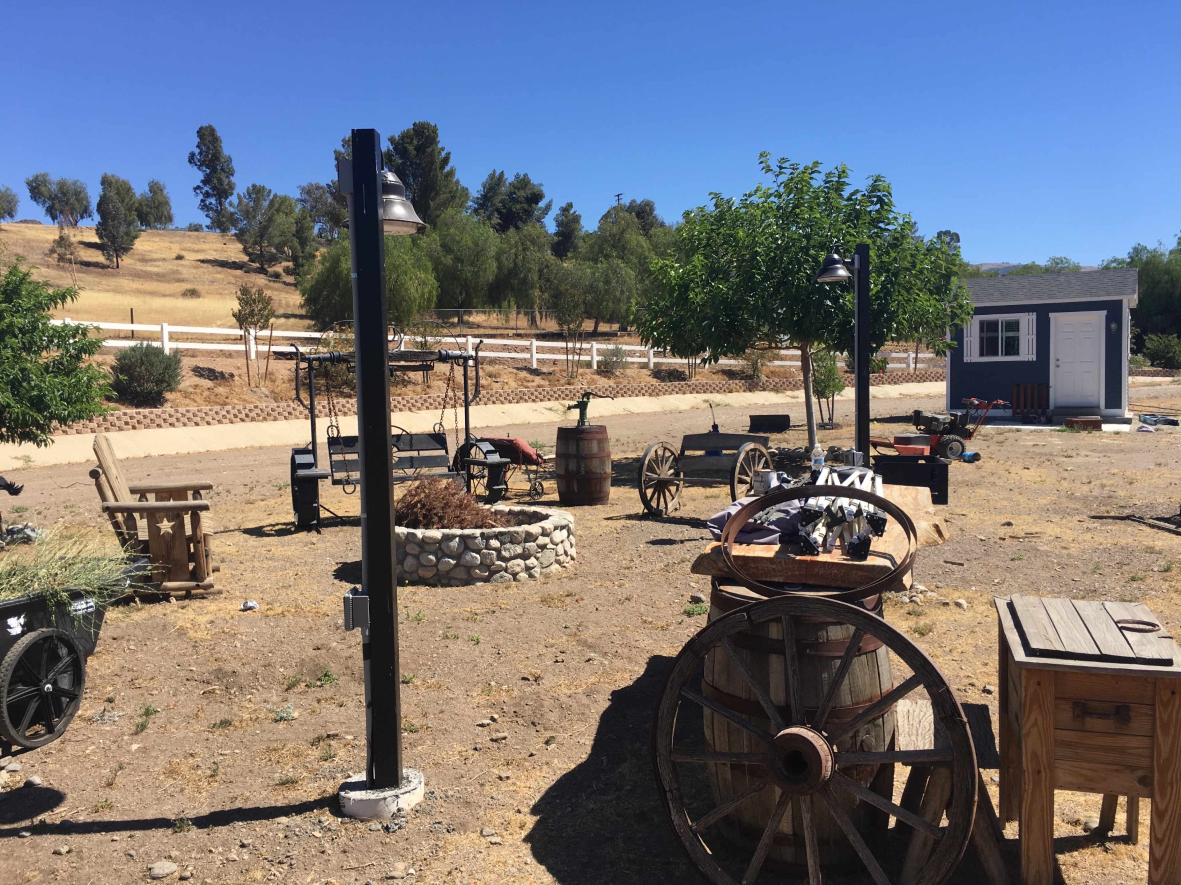 The scene features a rustic outdoor area with wooden carts, a fire pit, and a small shed surrounded by dry grass and trees.