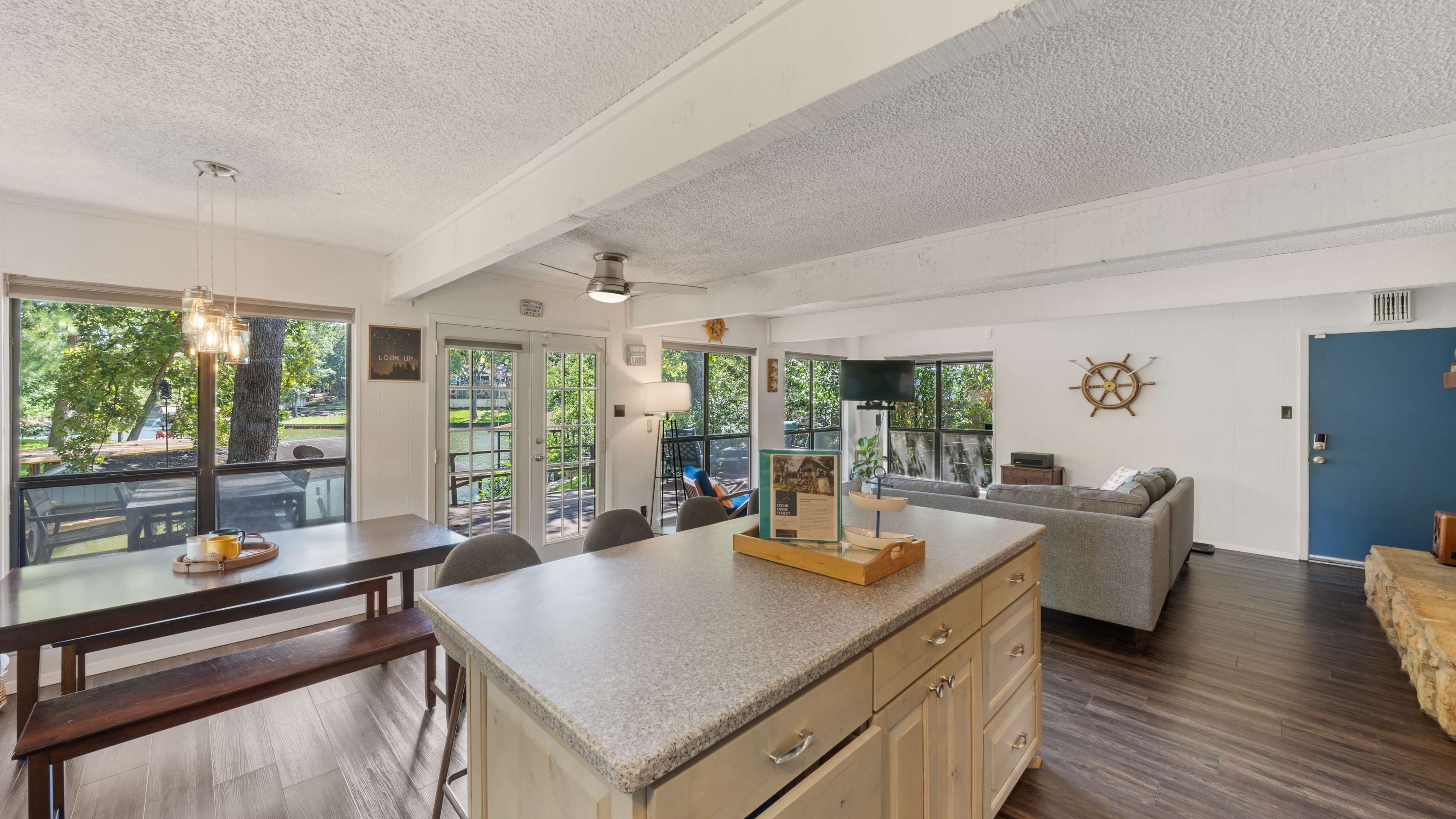 An open living space features a kitchen island, dining table, and a seating area with large windows that overlook a patio and outdoor greenery.