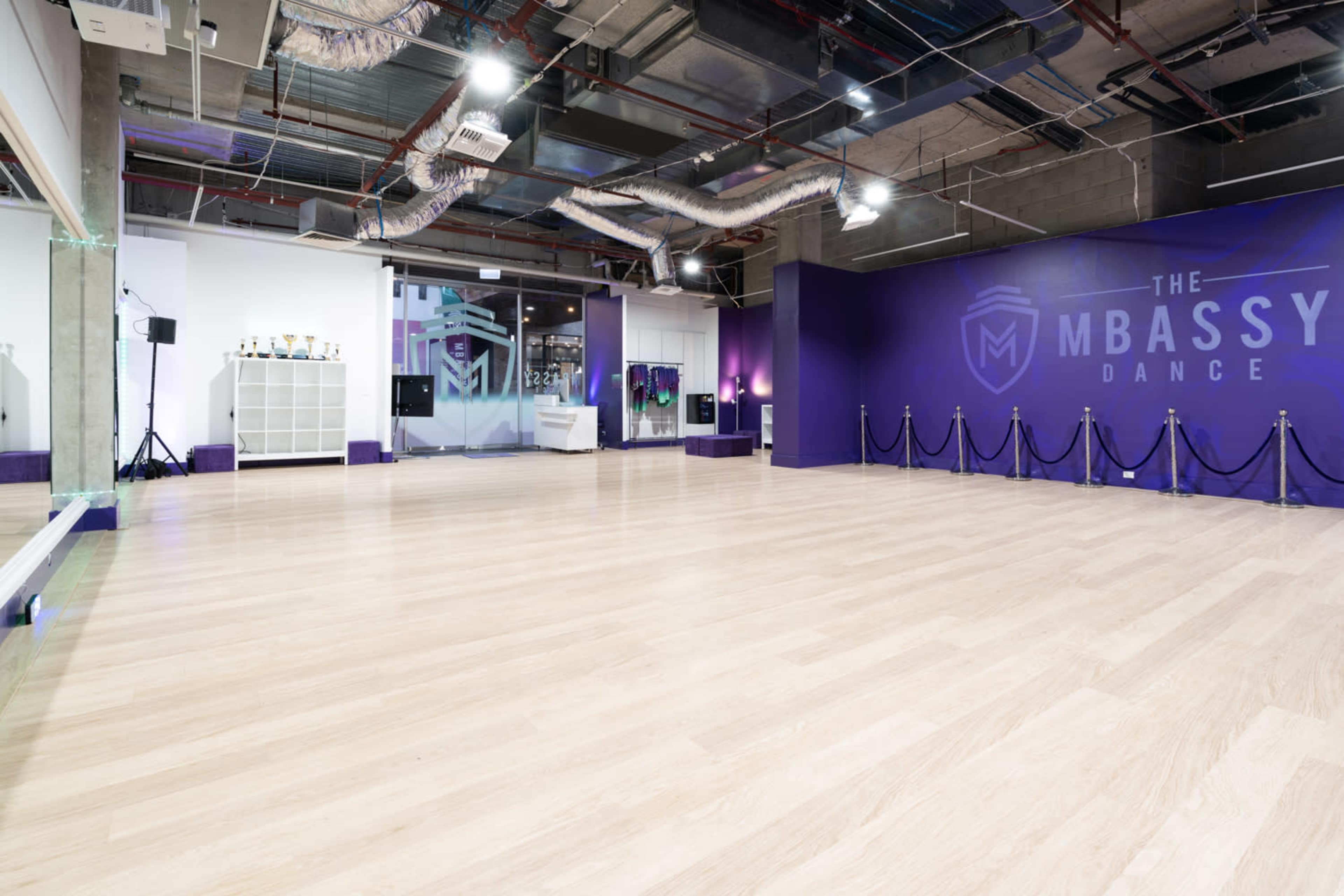 A spacious dance studio featuring a light wood floor, purple walls, and a minimalistic setup with a mirrored wall and trophies displayed.