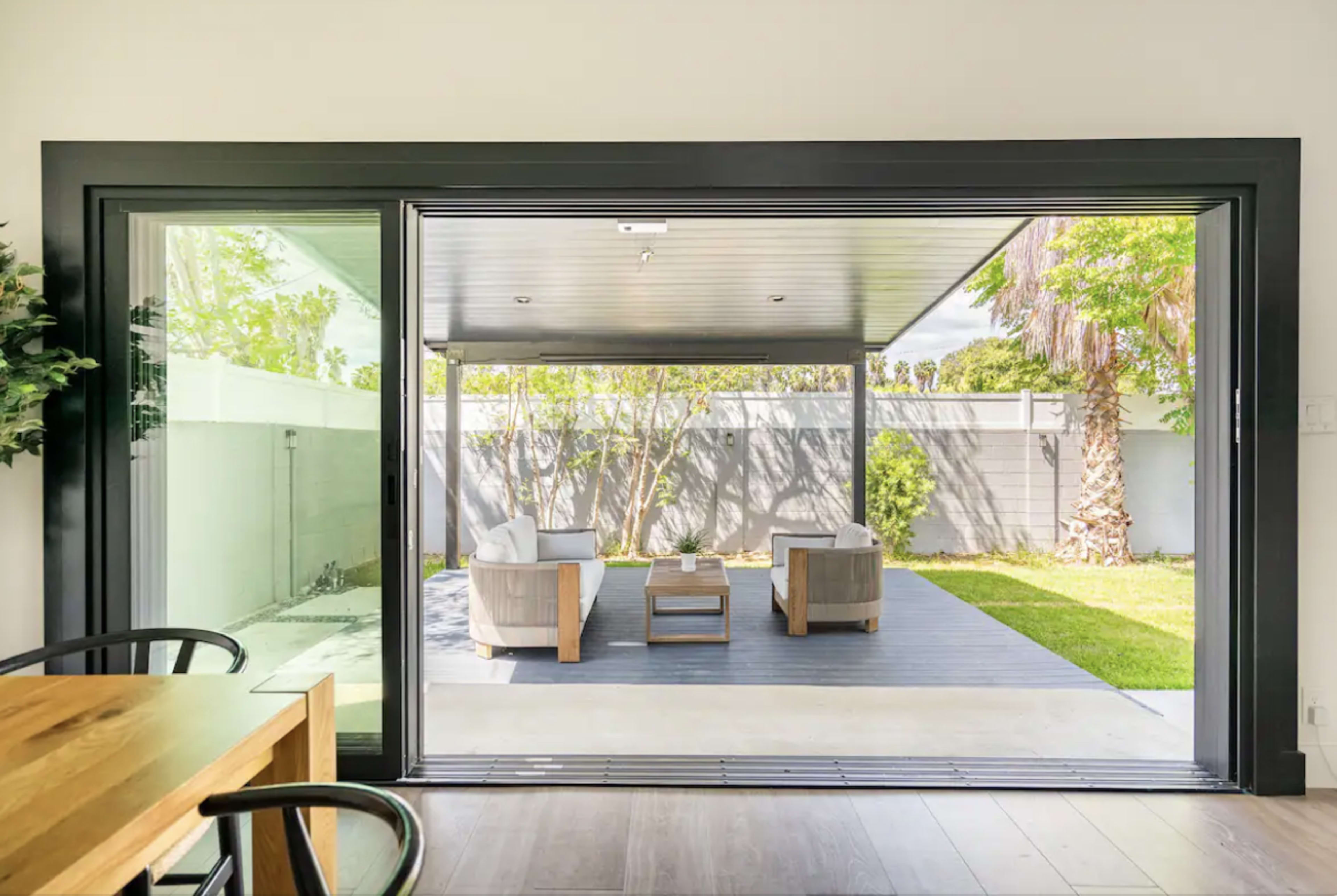 A modern indoor space opens to a patio featuring two armchairs, a coffee table, and a landscaped yard.