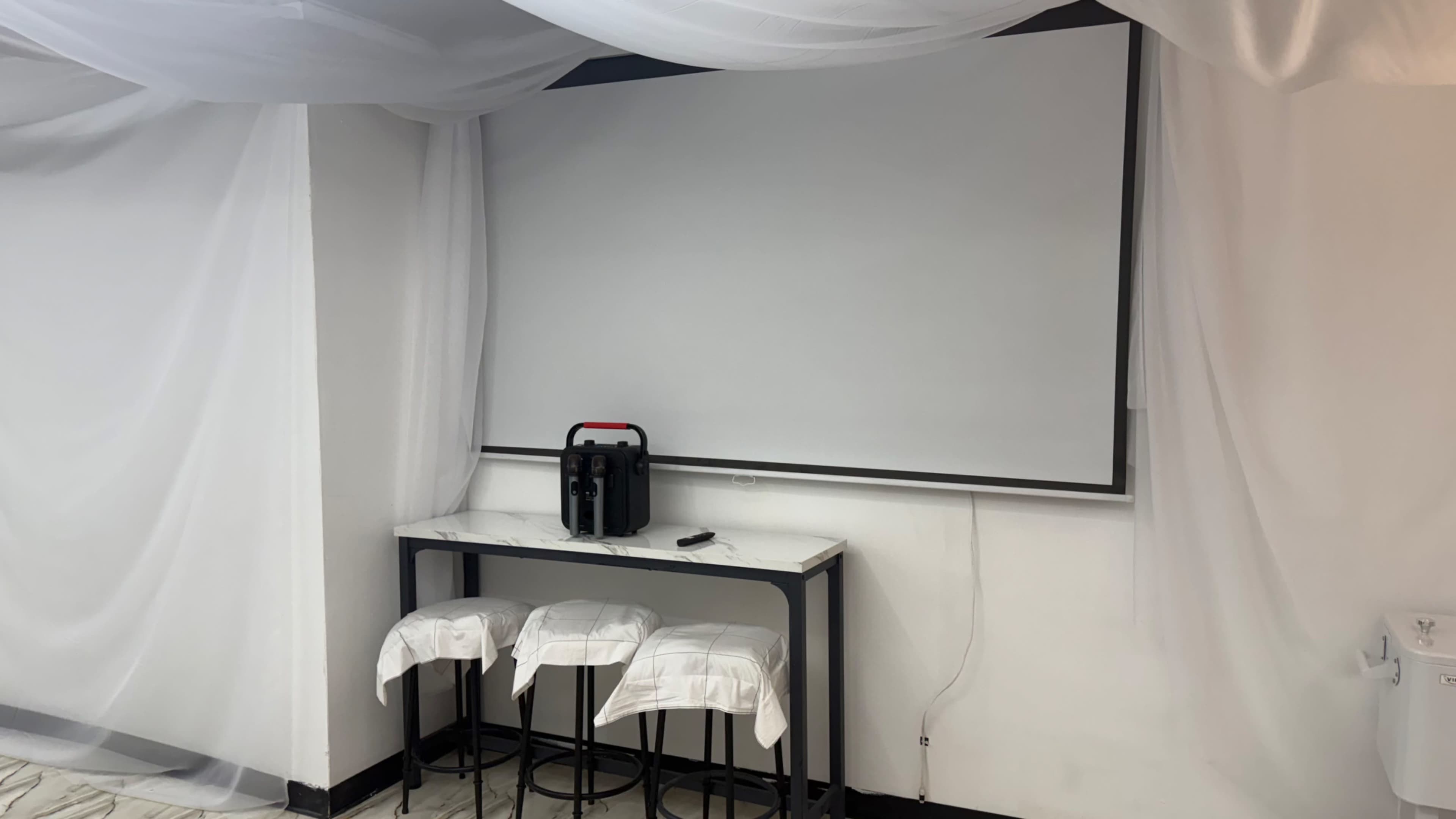 A minimalist room features a blank projection screen above a black table and three white bar stools draped with cloth.