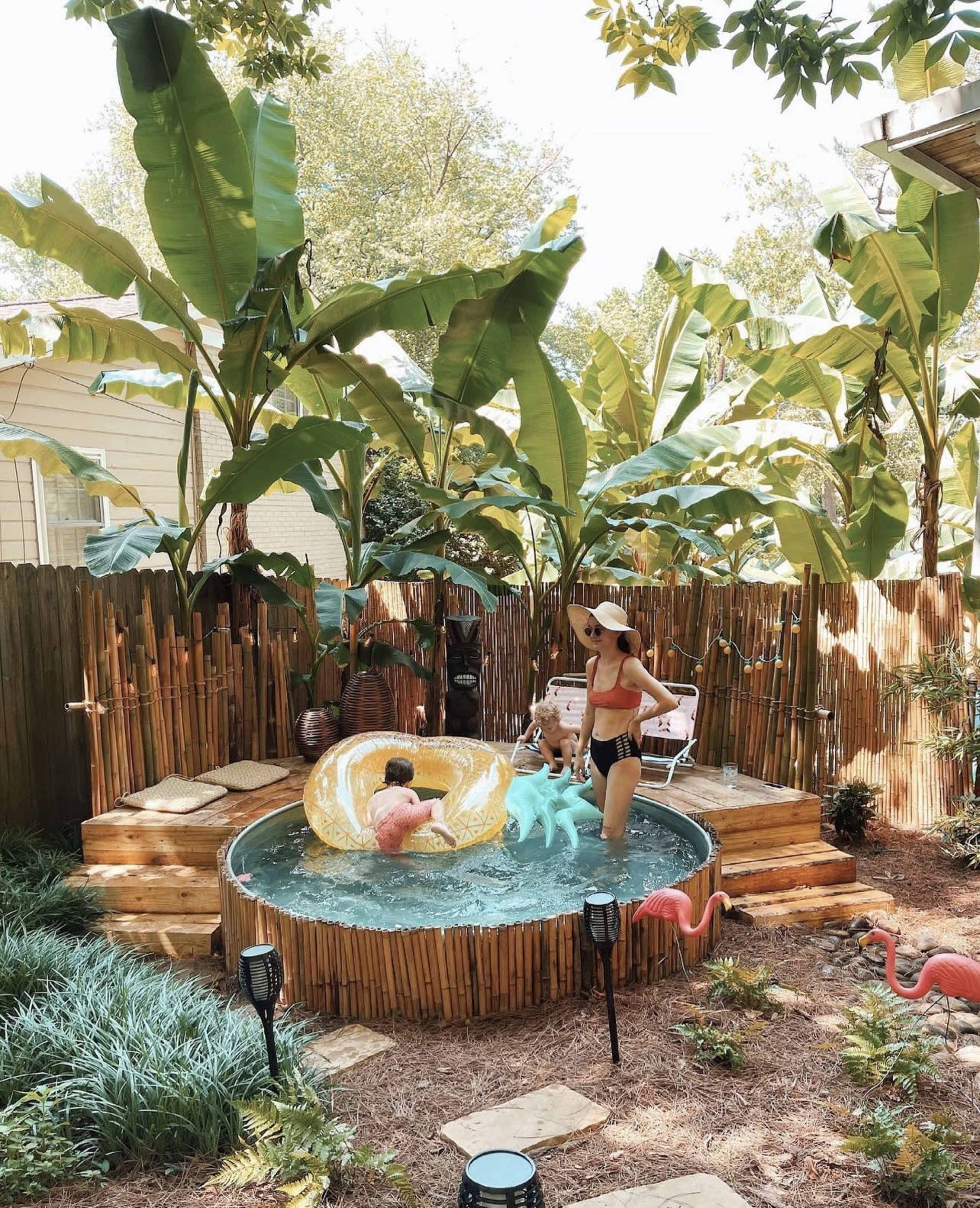 A boy plays in a small pool while a woman stands nearby in a backyard surrounded by tall plants and decorative flamingos.