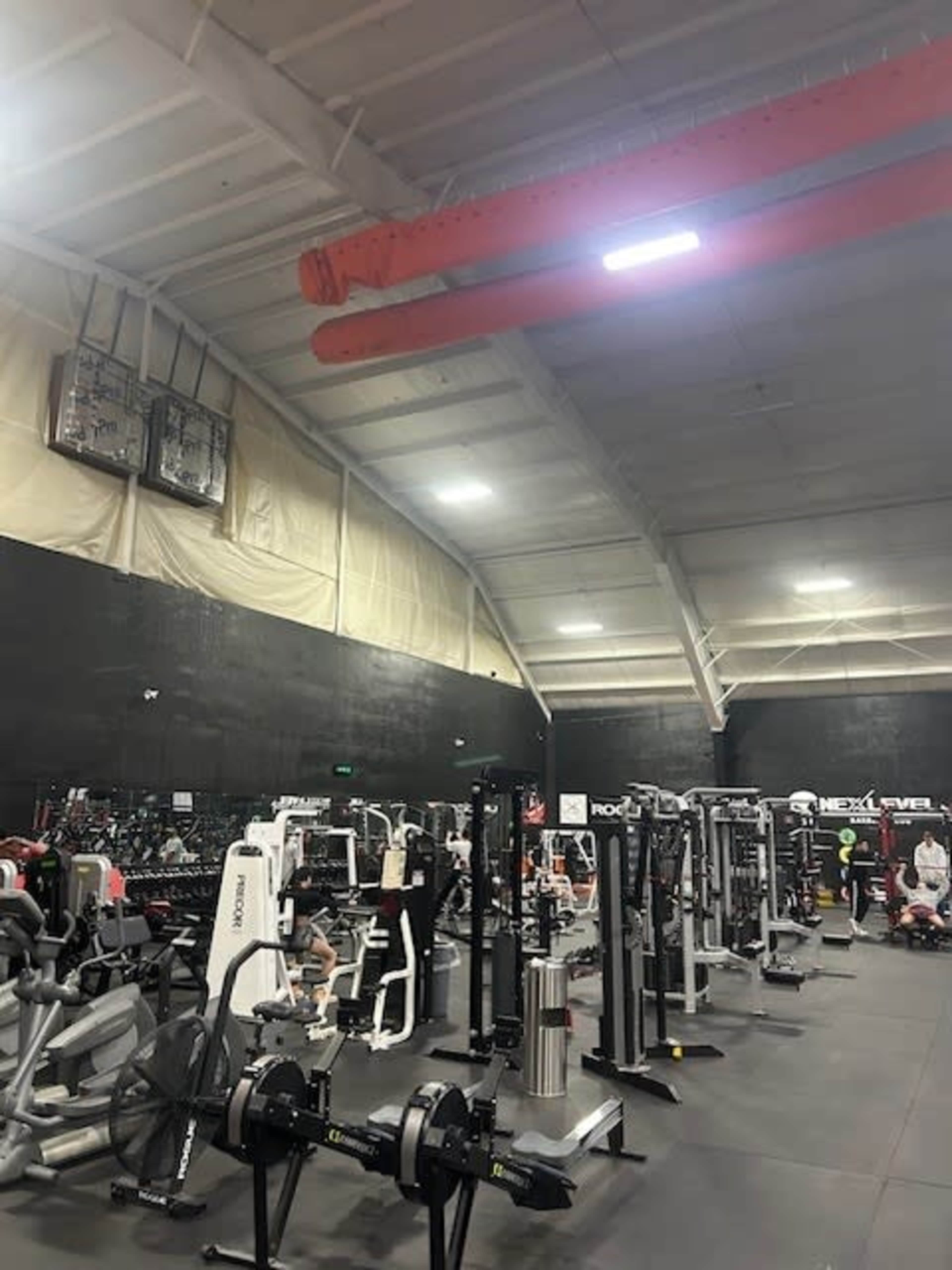 The image shows a spacious gym filled with various exercise machines and equipment under a high ceiling with exposed beams.