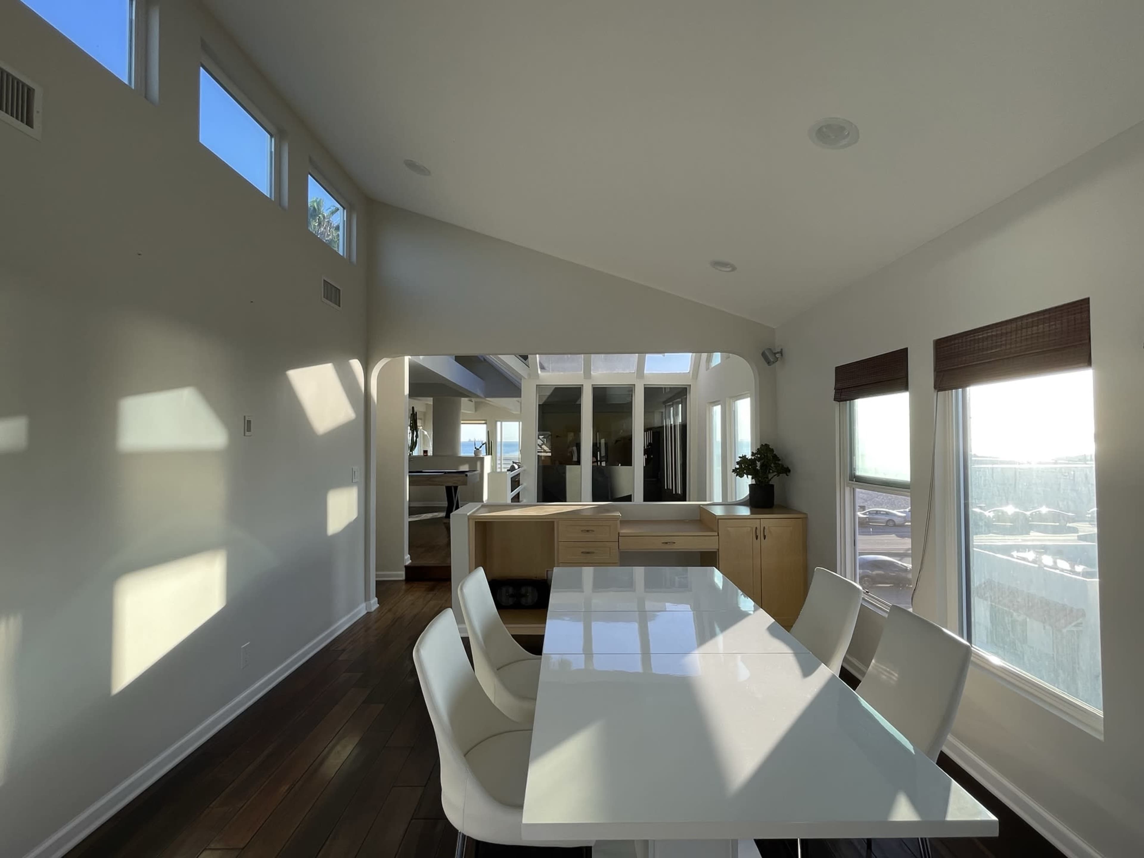 A bright dining area features a large white table surrounded by chairs, with large windows allowing natural light to fill the space.