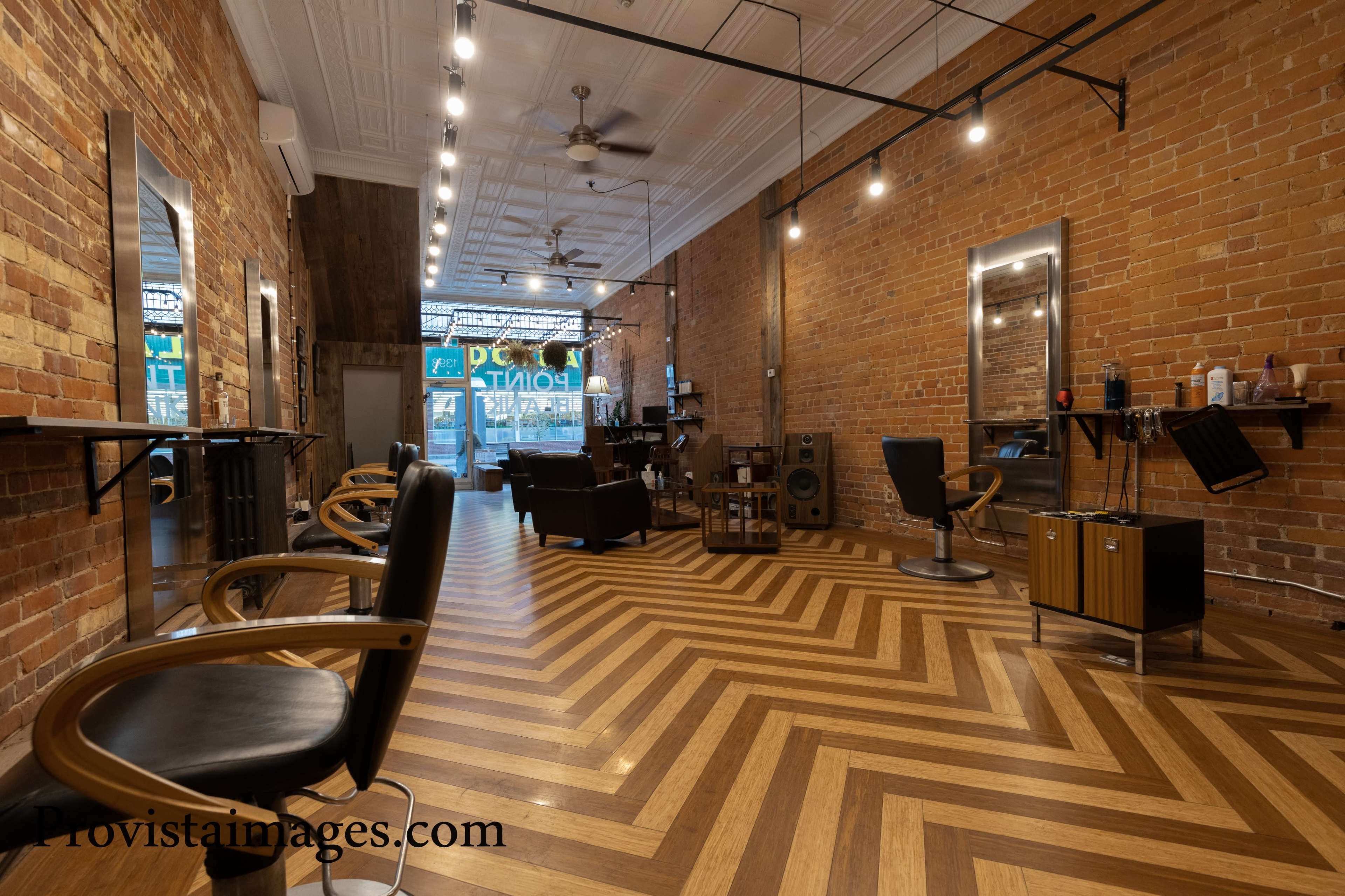An interior of a barbershop featuring exposed brick walls, wooden flooring in a herringbone pattern, and various styling stations with mirrors.