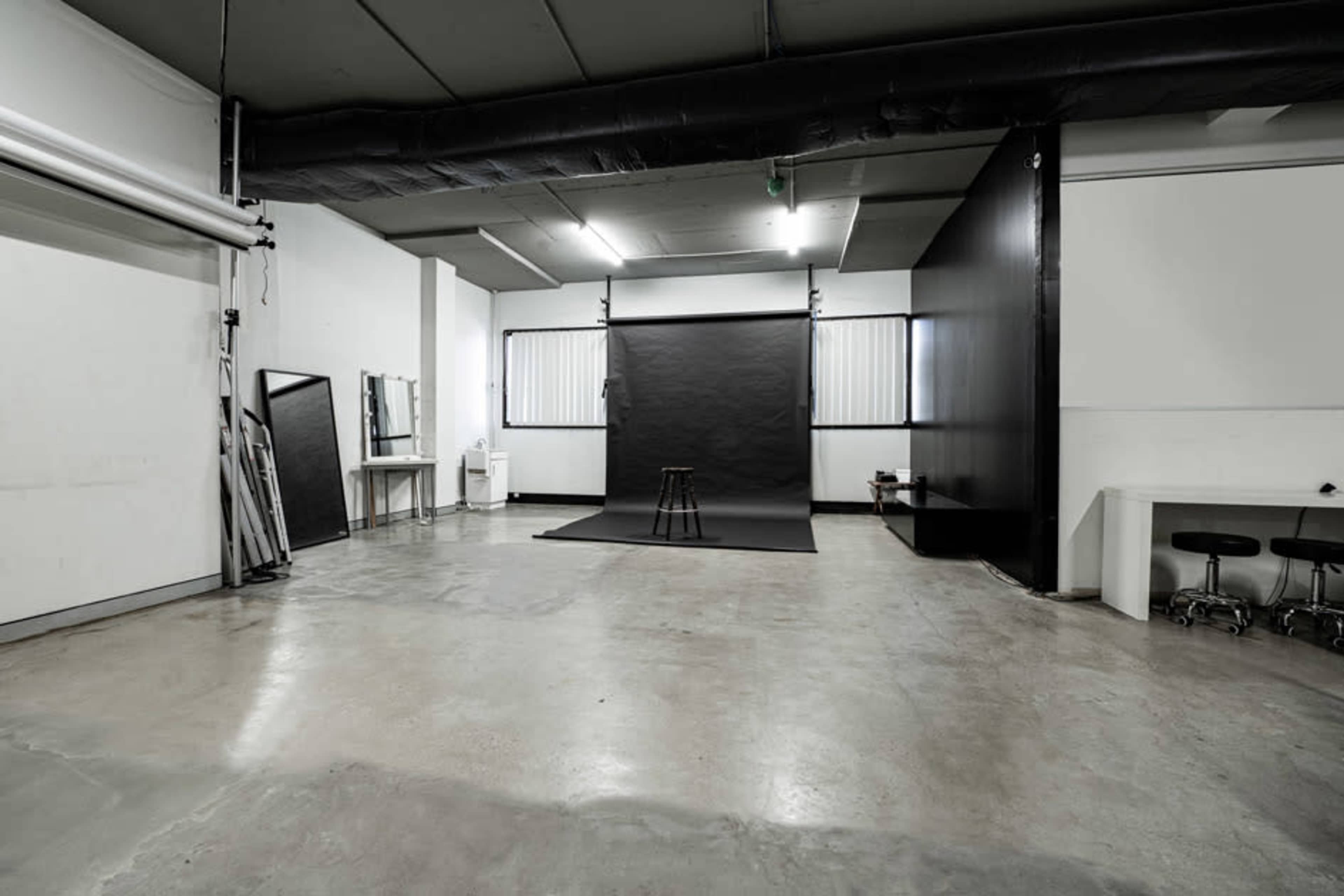 A spacious, empty photography studio with a black backdrop, a stool, and mirrored equipment against white walls.
