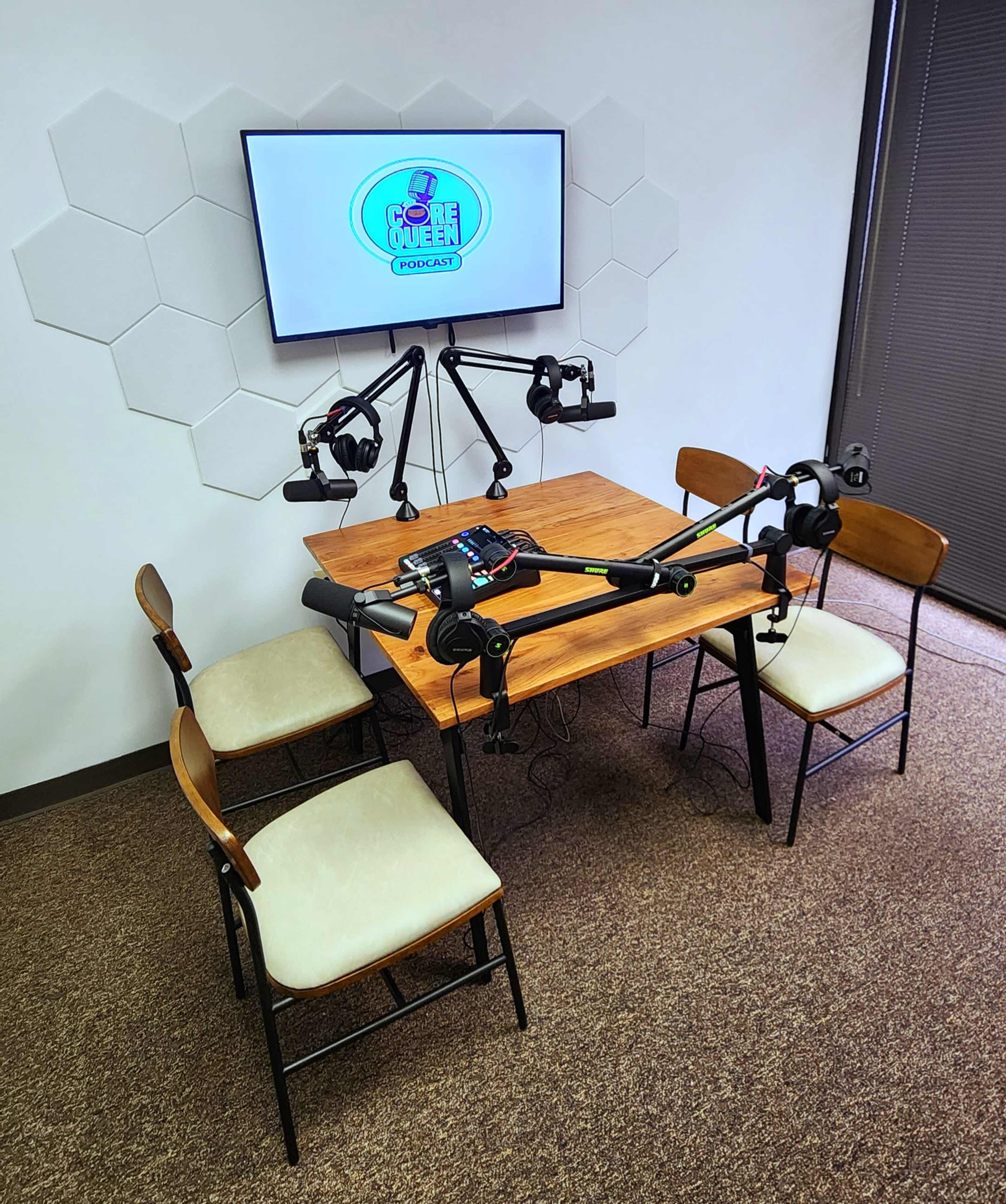 A small podcast recording setup featuring a wooden table with four chairs, equipped with microphone arms and a monitor displaying a podcast logo.