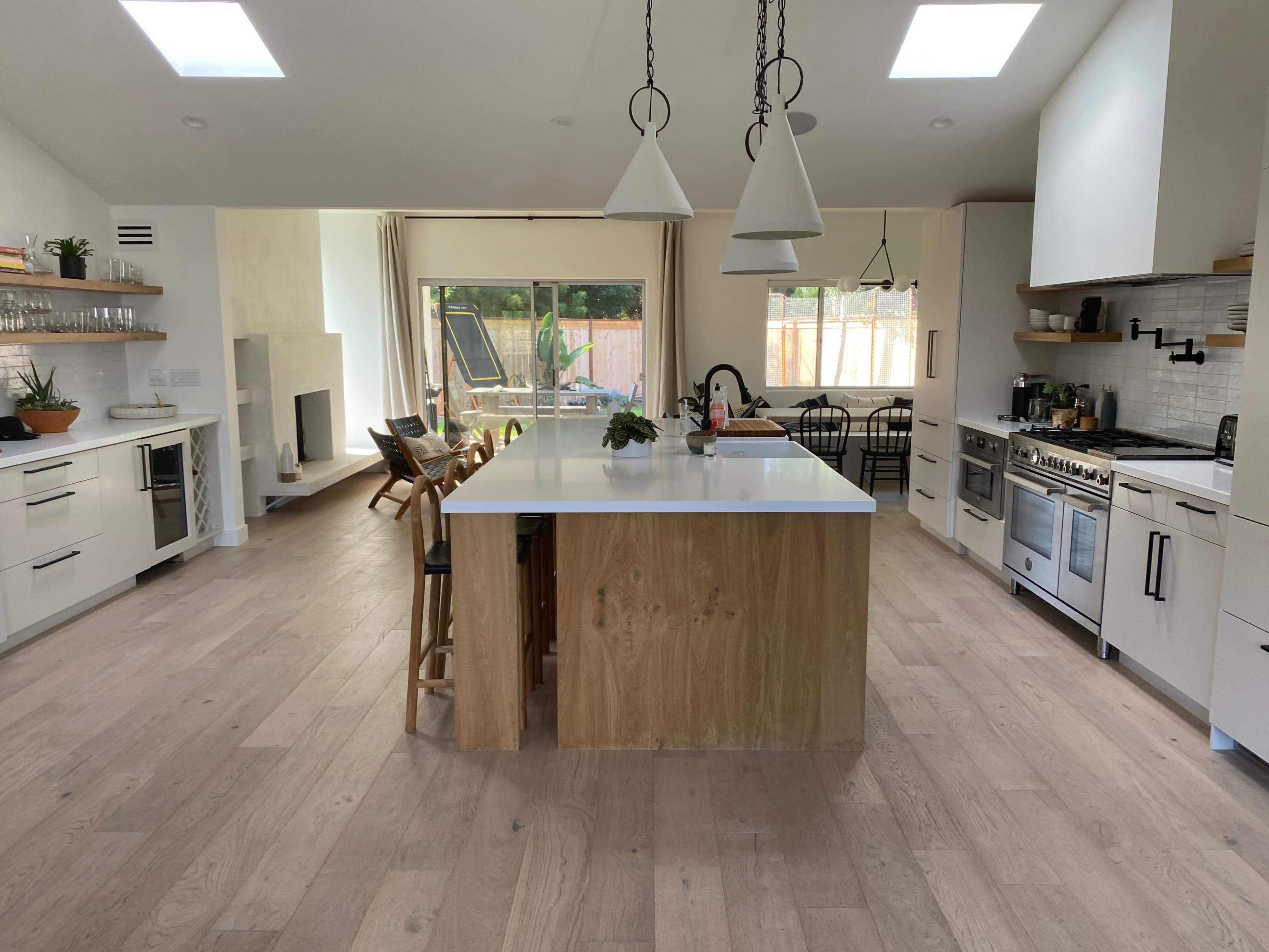 The image shows a modern kitchen featuring an island with wooden accents, white cabinetry, and a large window overlooking an outdoor area.
