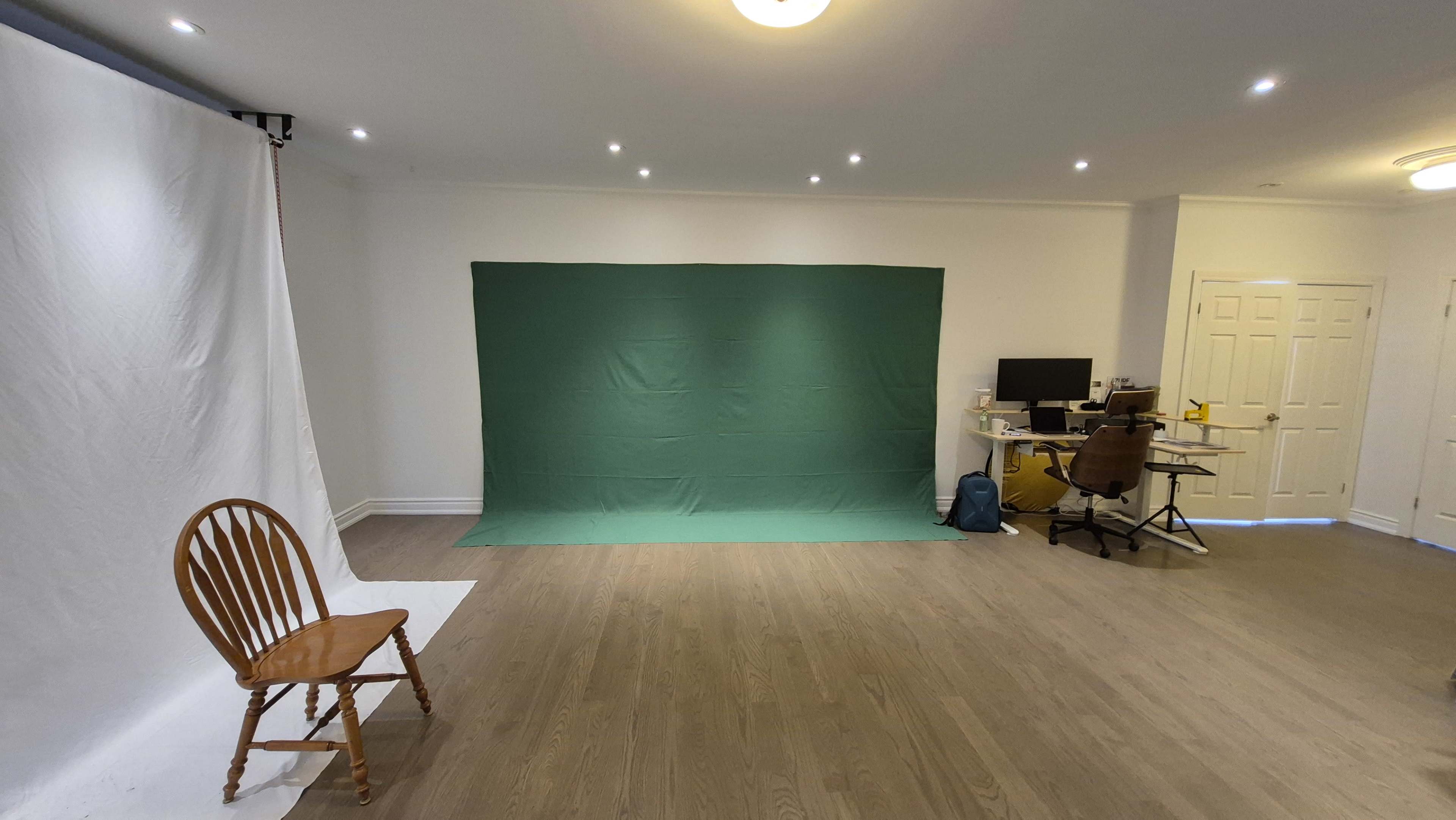 The image shows a spacious room with a green backdrop set against a plain wall, a white backdrop to the left, a wooden chair, and a desk with a computer in the corner.