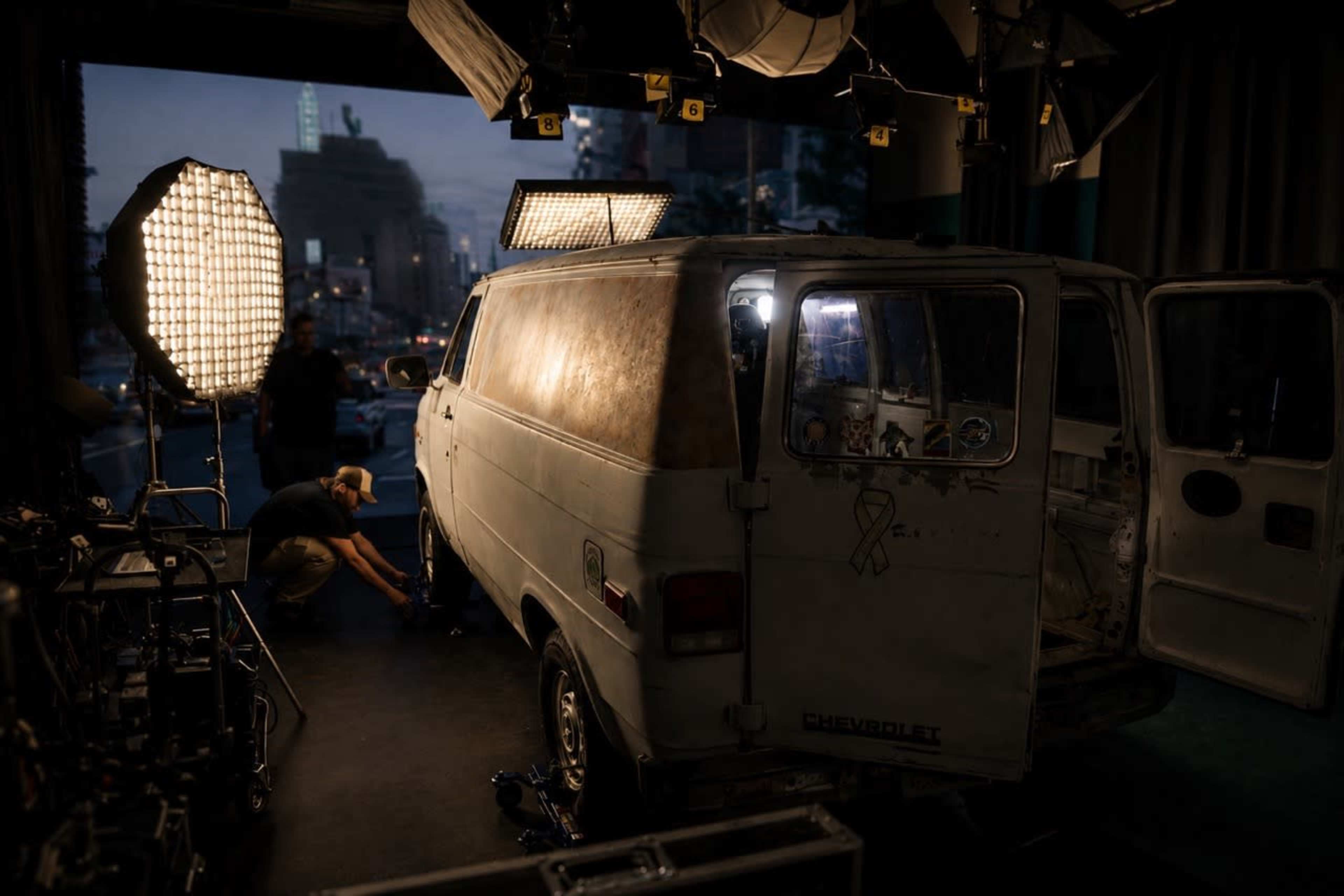 A man is working on a vintage Chevrolet van positioned in a dimly lit studio, surrounded by studio lighting equipment.